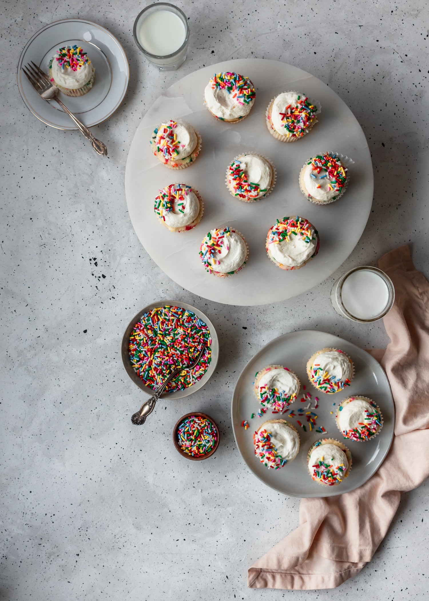 An overhead shot of sprinkle cakes on various white plates, surrounded by glasses of milk and bowls of sprinkles on a grey table.