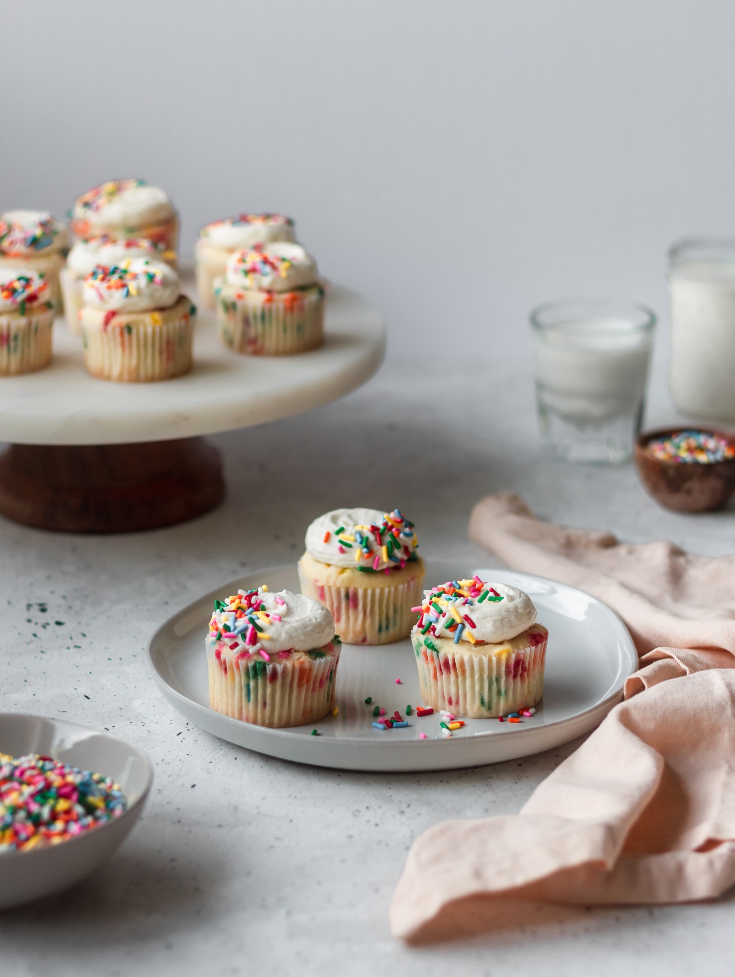 A straight-on shot of sprinkle cupcakes on a white plate on a grey table. In the background is a cake stand with more cupcakes, a couple glasses of milk, and some bowls of sprinkles.