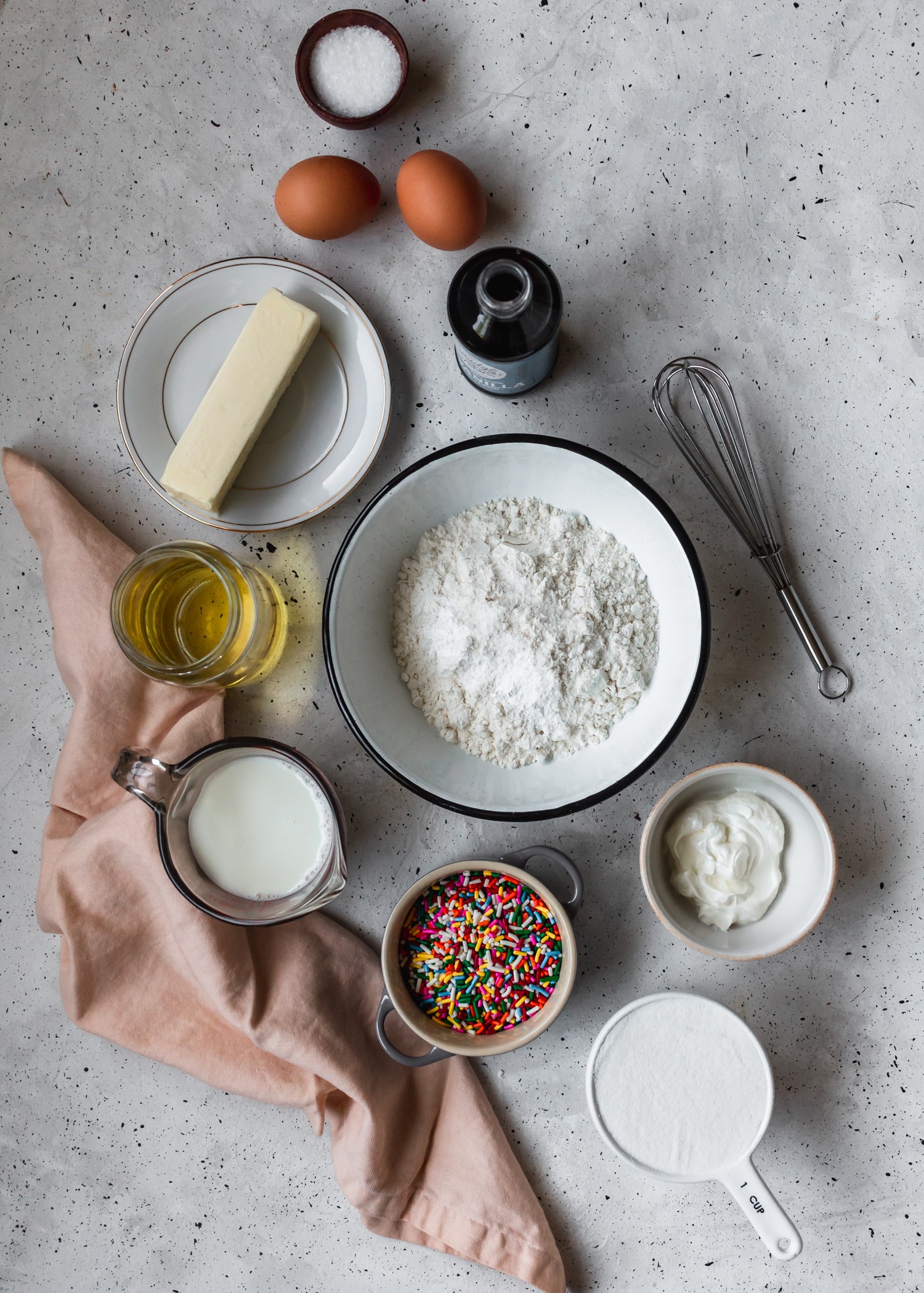 An overhead shot of cake ingredients with a pink linen on a grey speckled table.