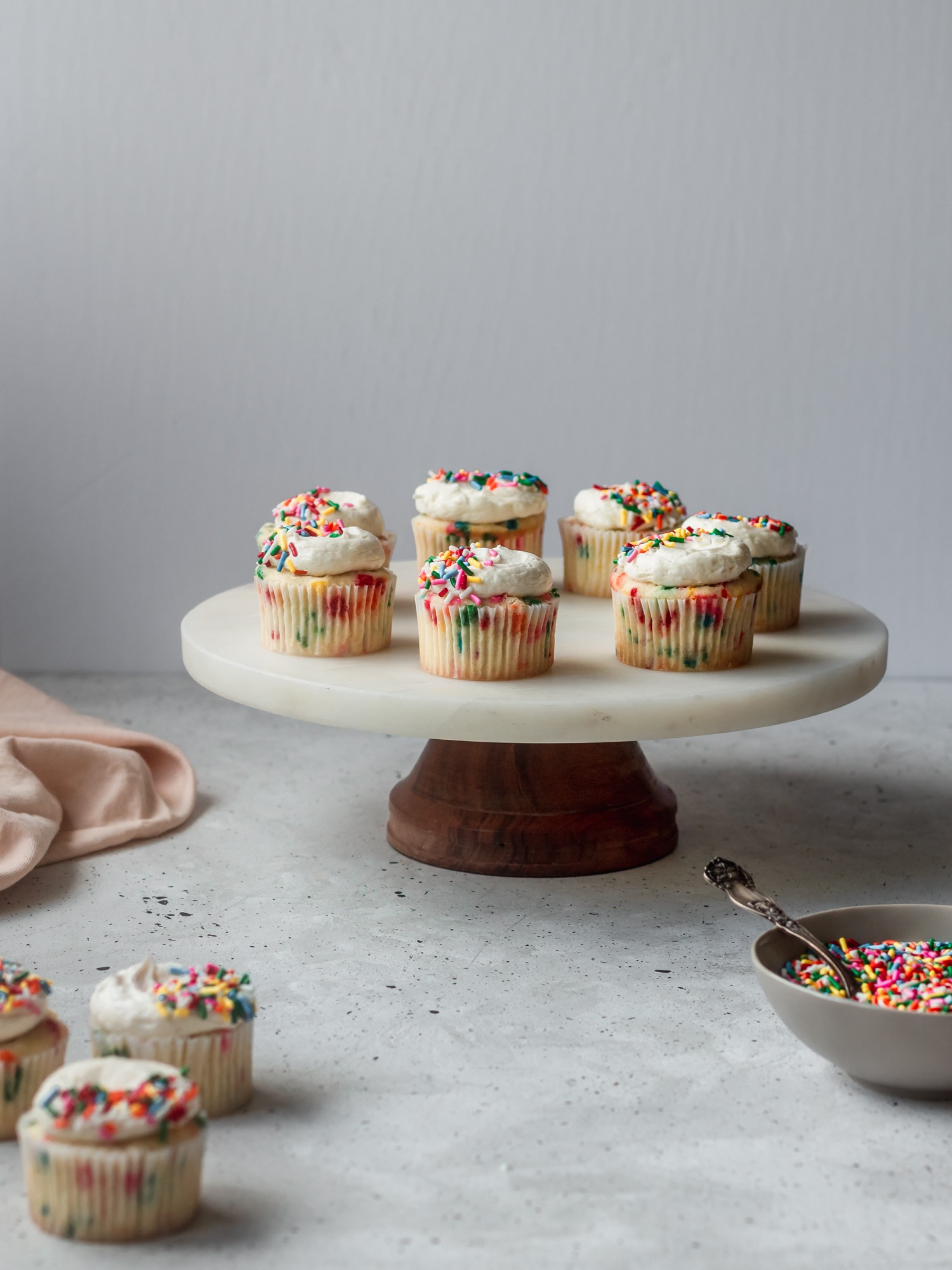 A marble cake stand with funfetti cupcakes on top next to a pink linen, more cupcakes, and a bowl of sprinkles on a grey table.