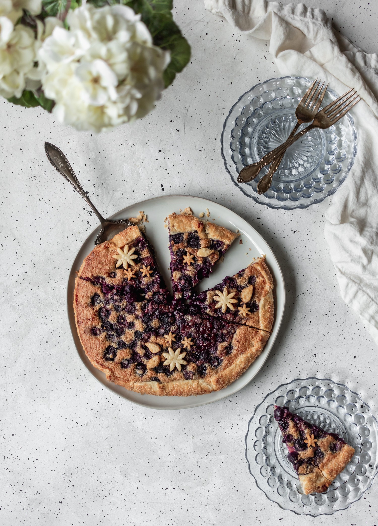 An overhead shot of a sliced blueberry galette on a grey background, surrounded by clear blue class plates, a white linen, and white hydrangeas.