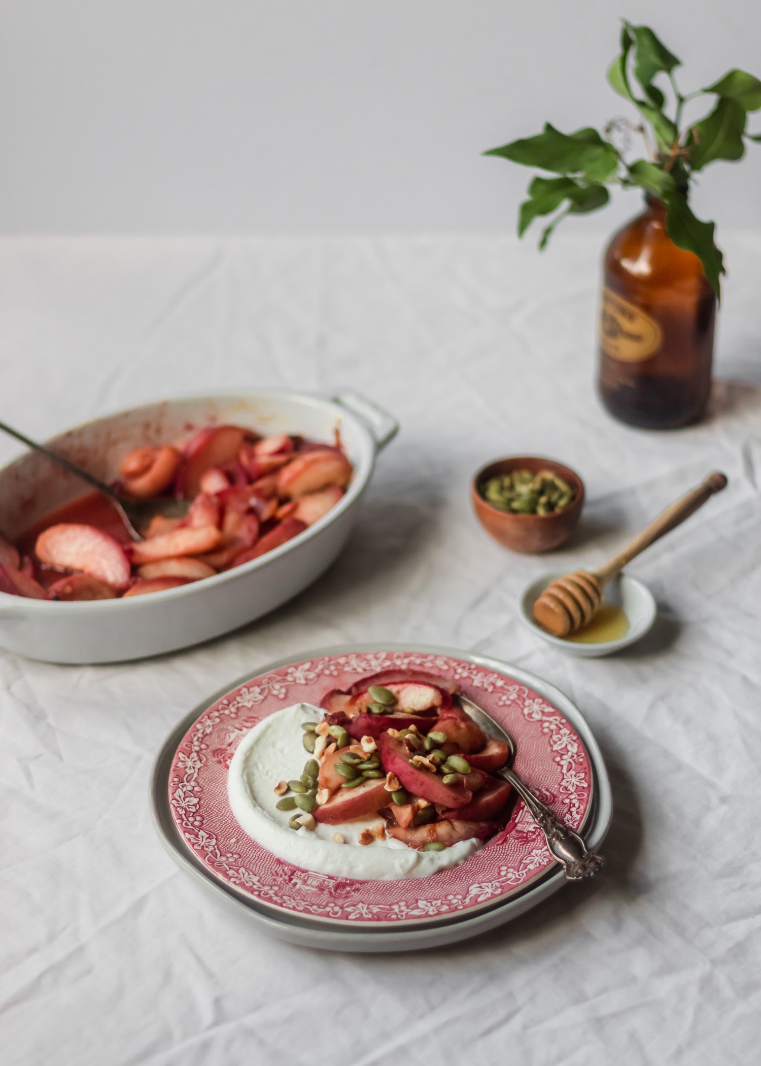 A side view of a red plate with roasted peaches on a white table cloth, with a roasting pan, honey, pumpkin seeds, and a jar of greenery in the background.
