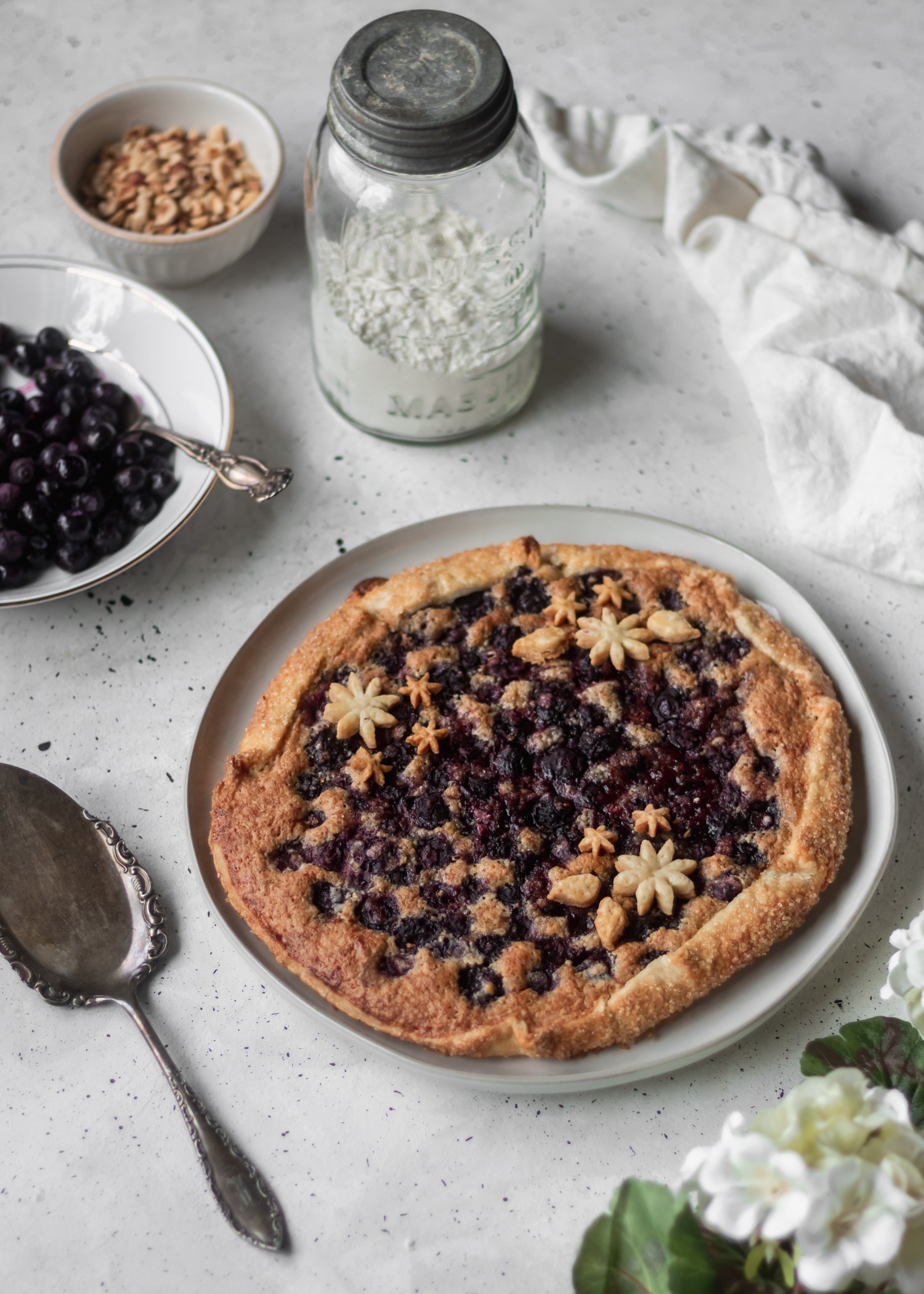 A side shot of a blueberry galette on a white plate sitting on a grey table surrounded by white hydrangeas, a pie knife, and pie ingredients.