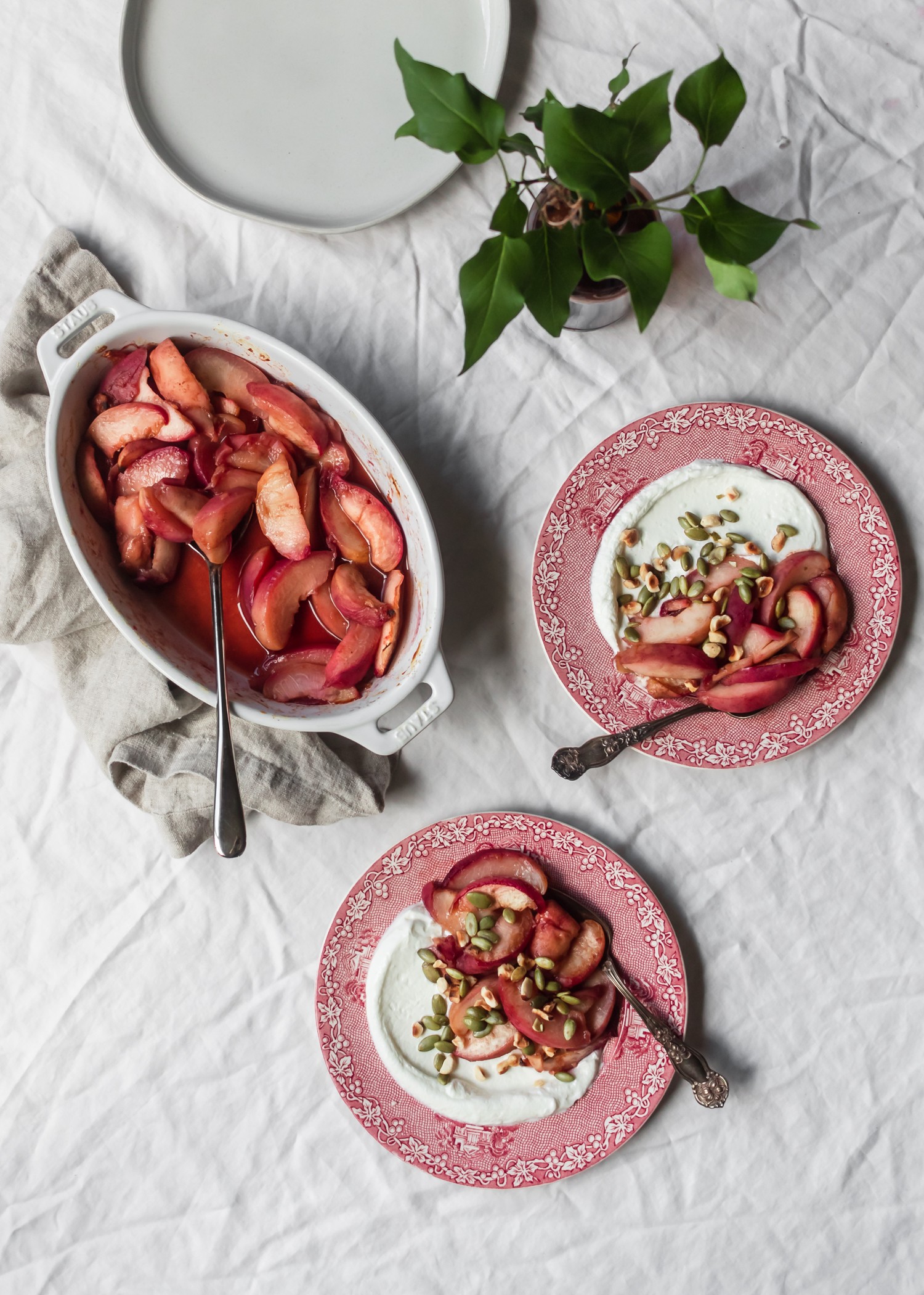 An overhead shot of two red plates with roasted peaches and yogurt on a white linen next to a roasting pan with more peaches and a vase of greenery.