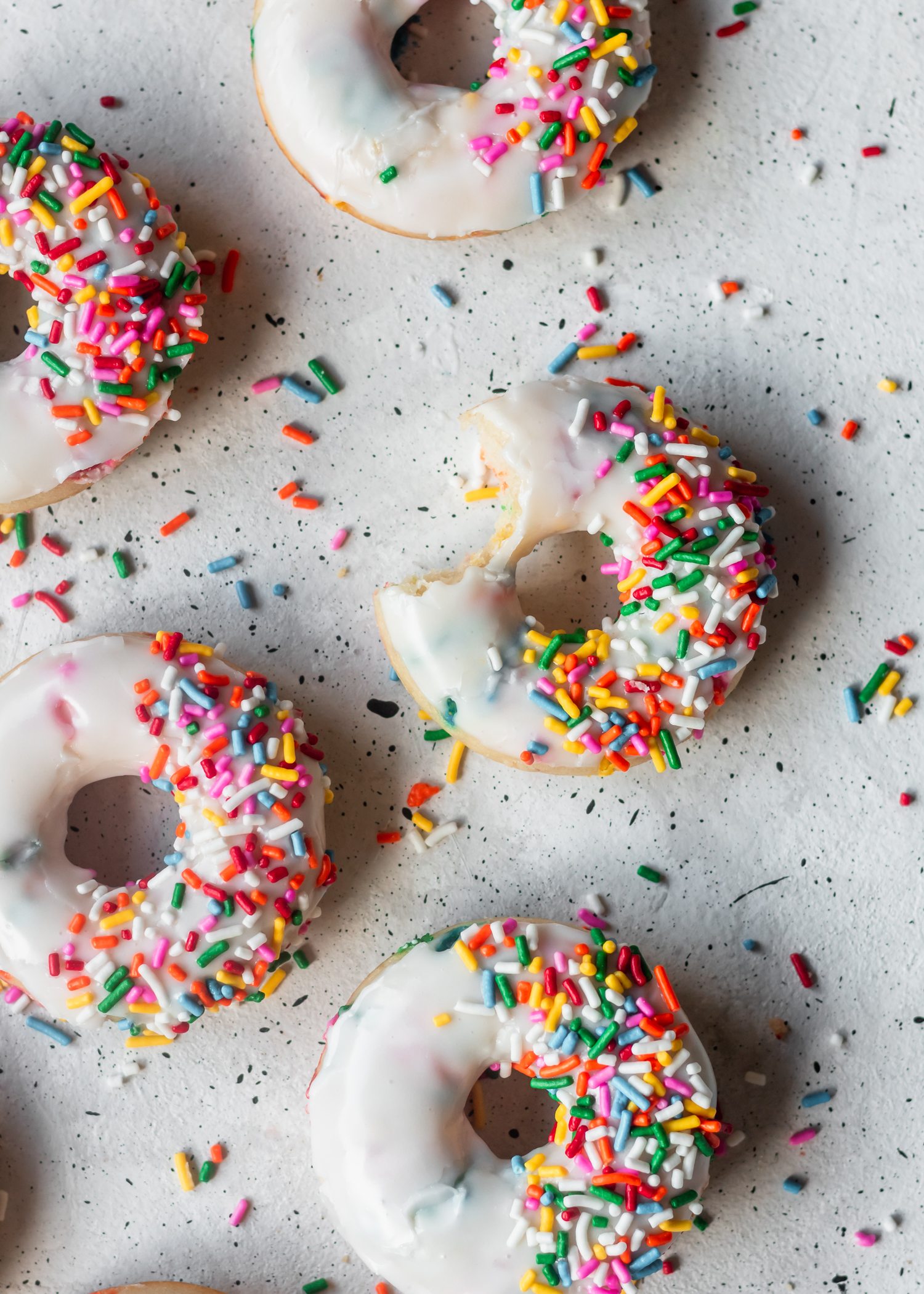 An overhead shot of a funfetti donut with a bite taken out, surrounded by other donuts on a white speckled background with rainbow sprinkles.