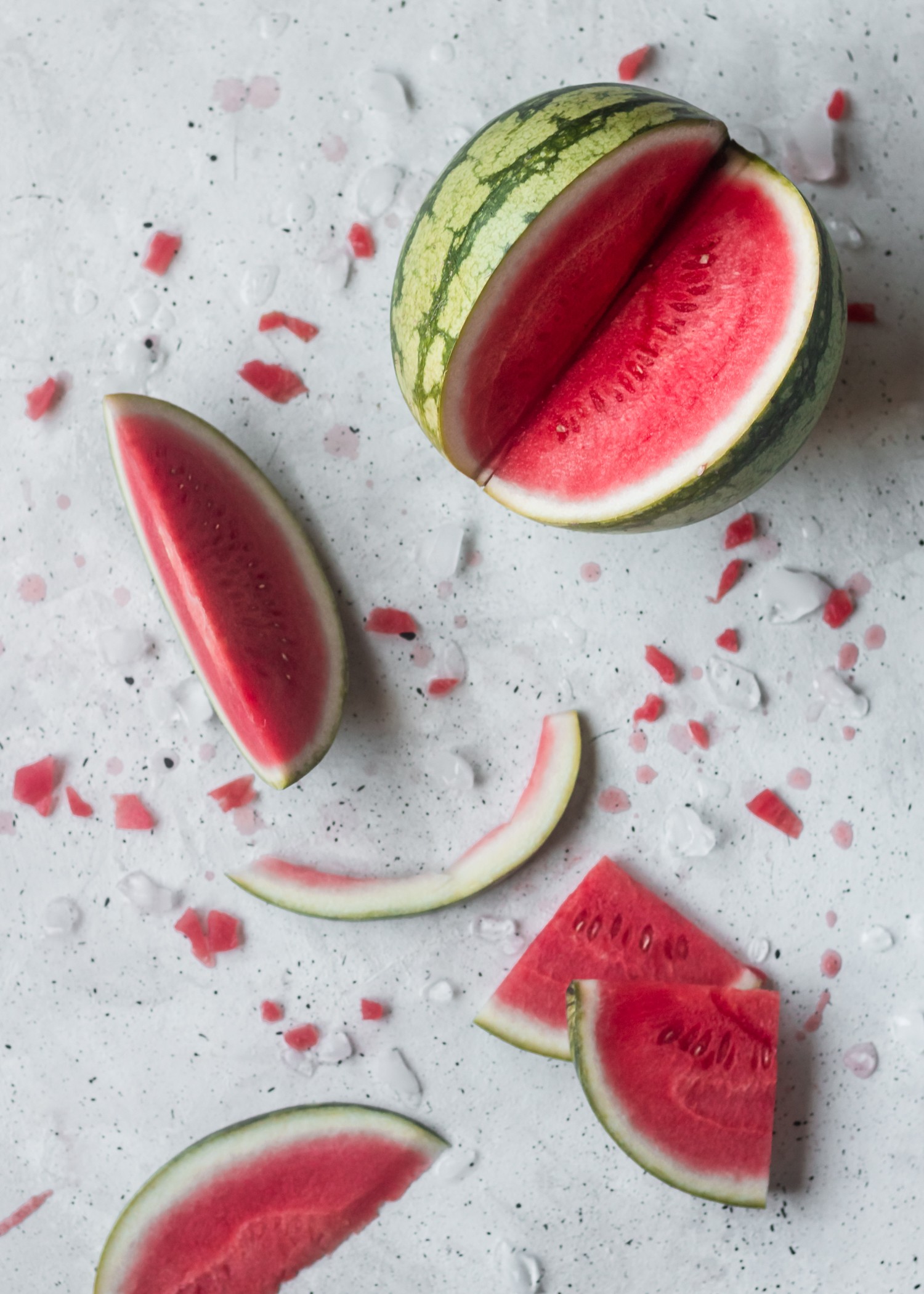 Overhead photo of a sliced watermelon on a grey speckled background, surrounded by watermelon slices, rind, and juice.
