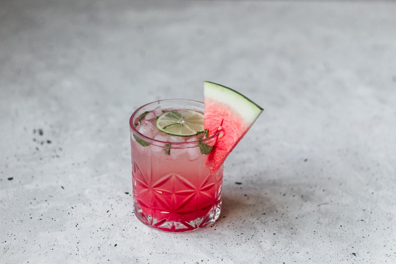 A side photo of a watermelon gin fizz garnished with a slice of melon on a grey speckled table.