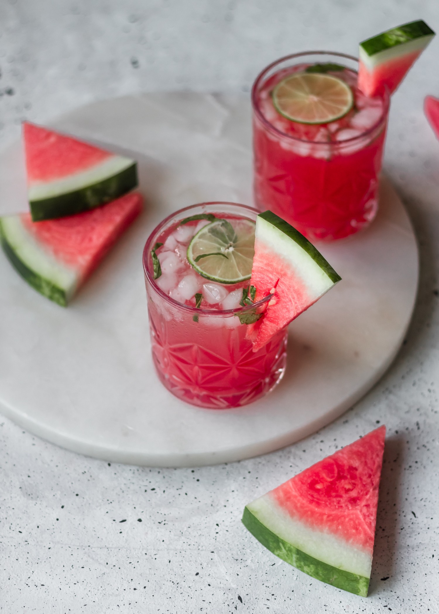 A side shot of a watermelon gin fizz on a white marble plate, next to another cocktail, surrounded by watermelon slices.
