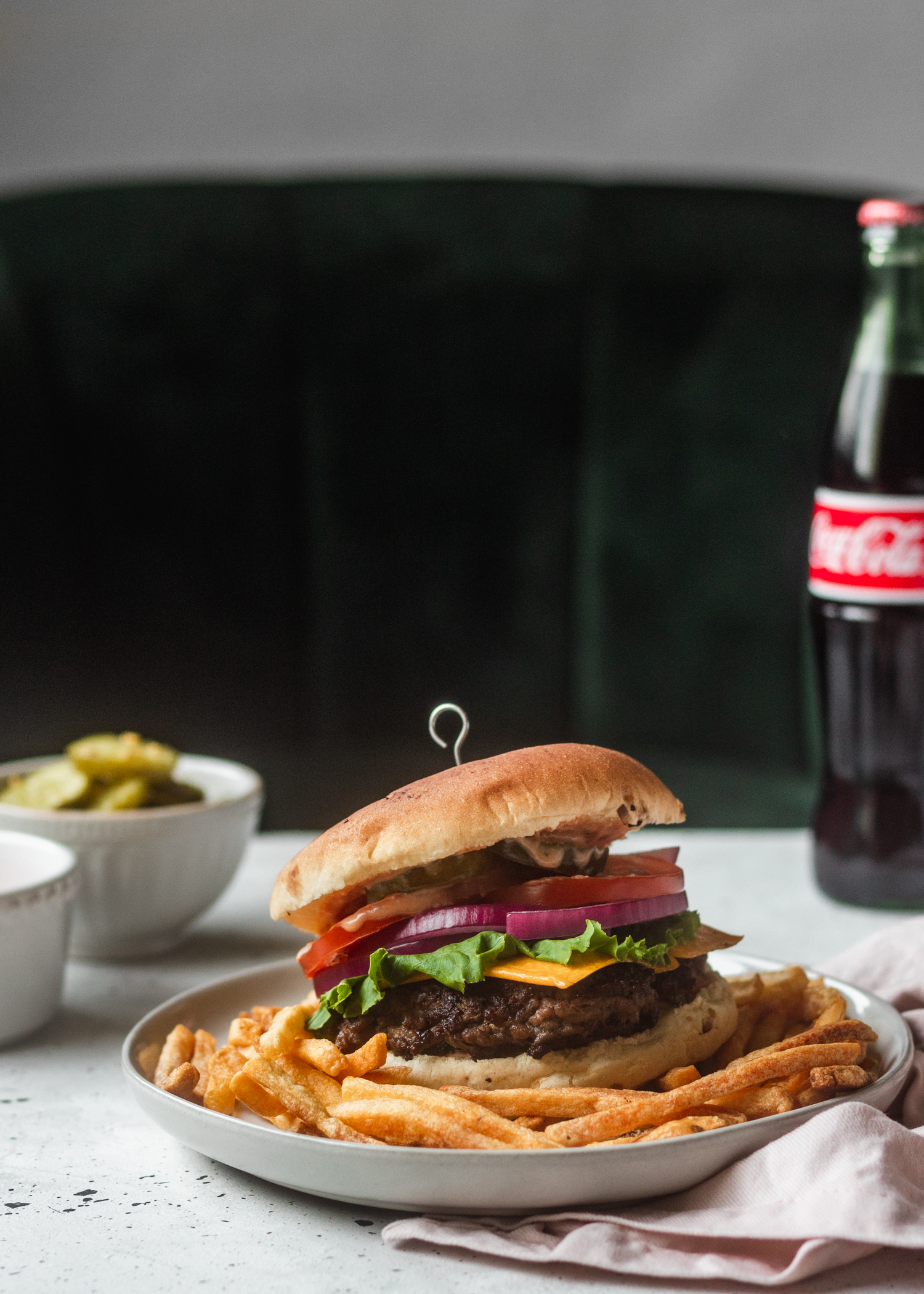 A close-up of a cheeseburger on a white plate with fries, sitting on a grey table next to a bottle of Coke, a pink linen, and a bowl of pickles with a green velvet booth in the background.