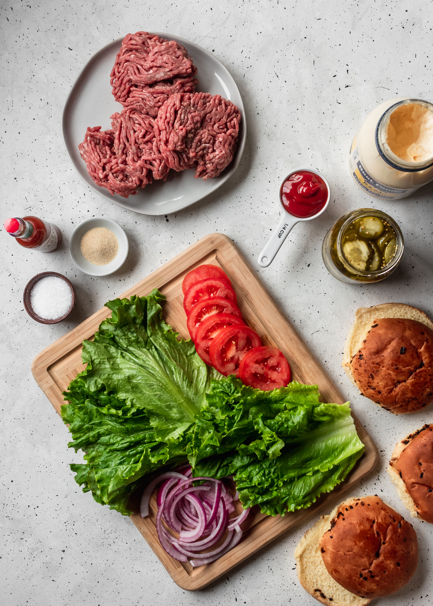 An overhead image of a wood board with veggies, a white plate of ground beef, buns, pickles, and condiments on a white speckled counter.