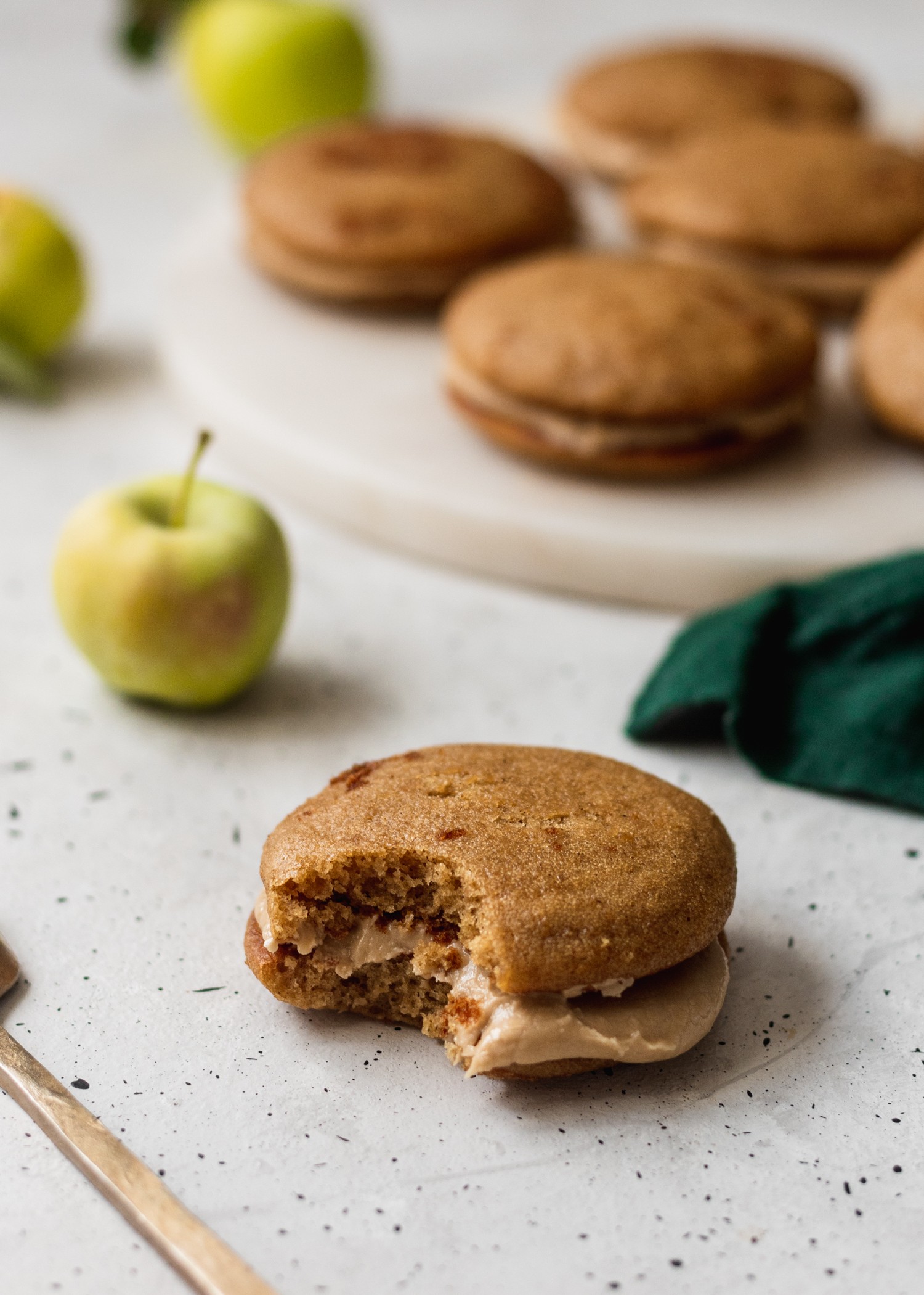 A close up shot of an apple cider whoopie pie with a bite taken out of it sitting on a white speckled table with a plate of apple cider whoopie pies, a green linen, and an apple in the background.