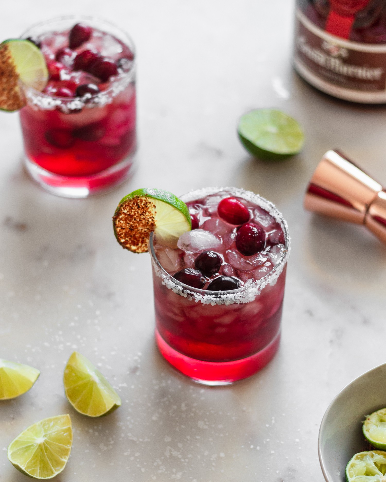 A 45 degree shot of a cranberry margarita on a white marble table surrounded by additional margaritas, a copper shot glass, a bottle of Grand Marnier, and limes.