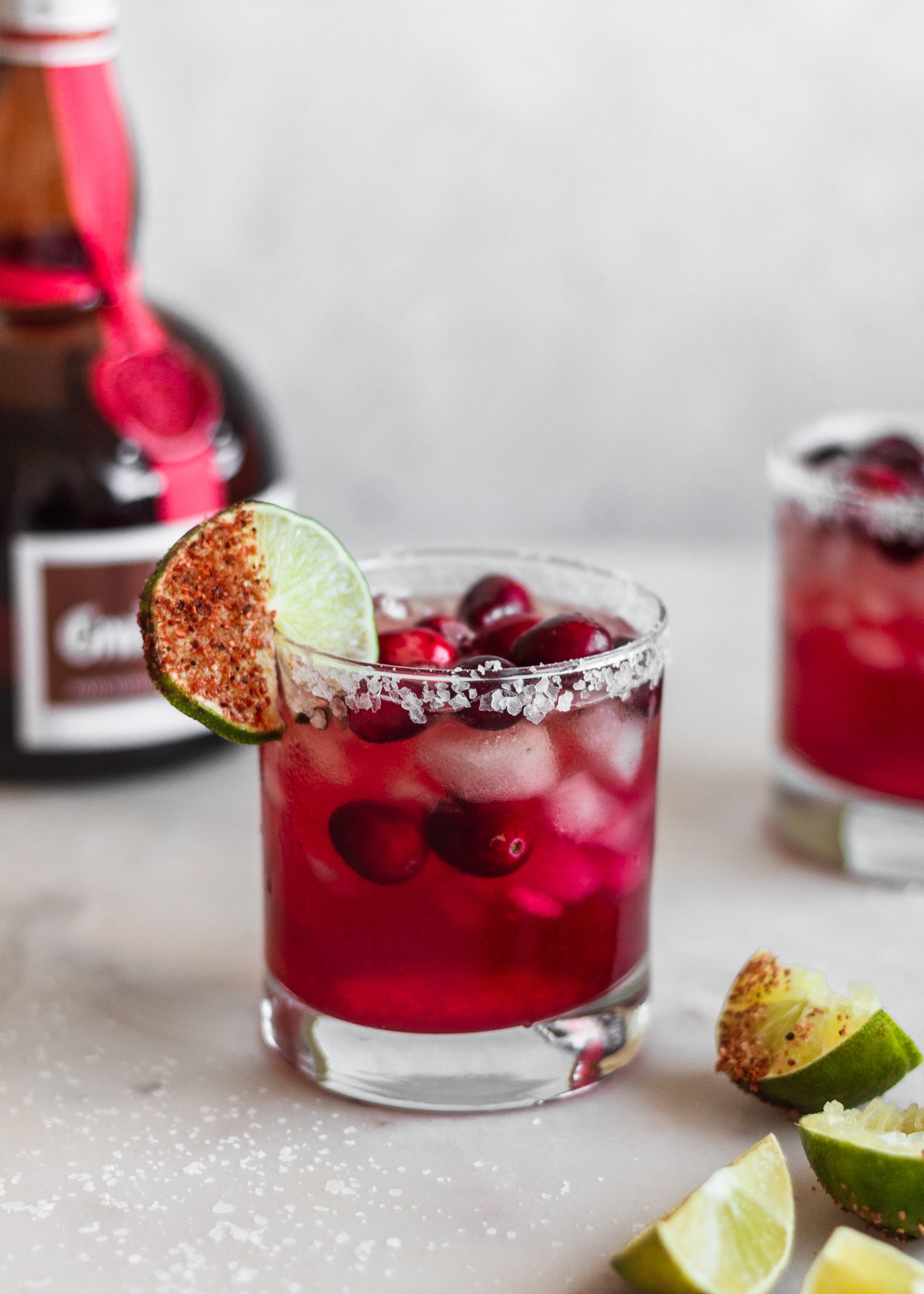 A close-up side shot of a cranberry margarita on a white marble table surrounded by limes, a bottle of Grand Marnier, and more cranberry margaritas.