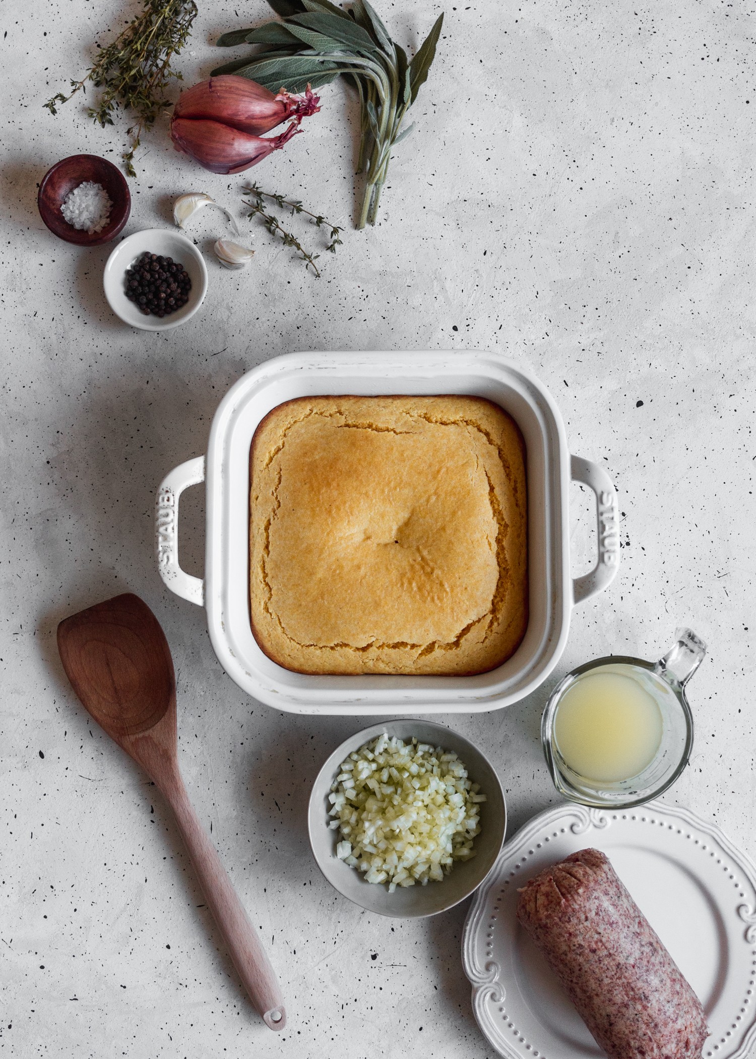 An overhead photo of a pan of cornbread surrounded by sausage, herbs, celery, chicken stock, and a wooden spatula on a grey table.