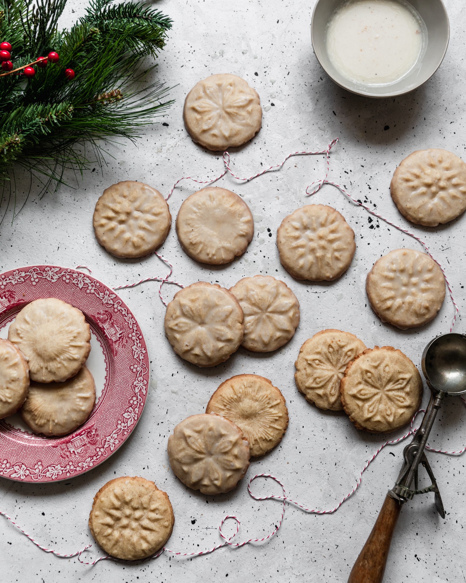 An overhead photo of stamped cookies on a grey counter surrounded by a plate of cookies, pine, and a cookie stamp.
