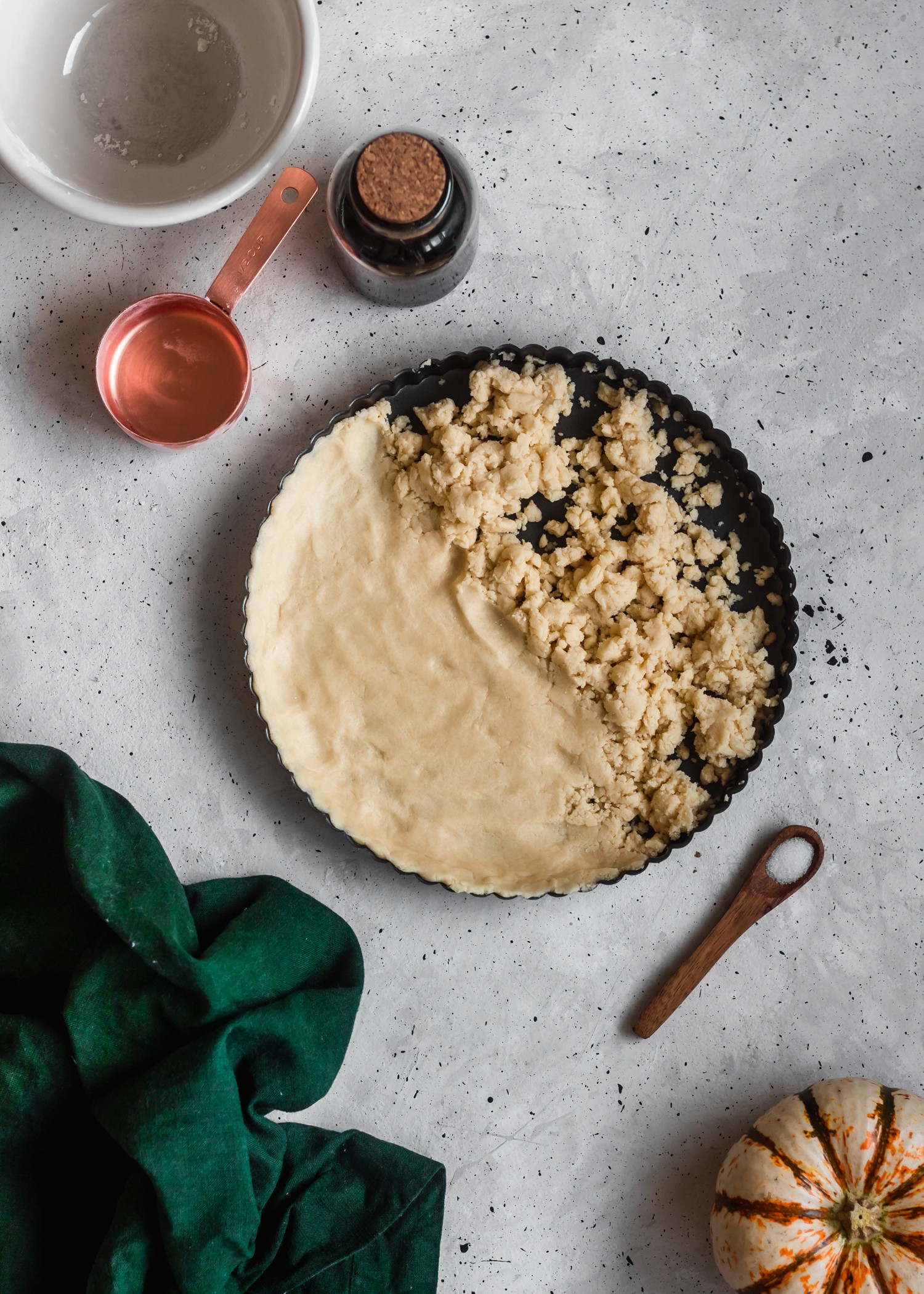 A bird's eye view of a shortbread crust in a black tart pan placed on a grey counter next to a green towel, wooden spoon, white and orange pumpkin, and copper measuring cup.
