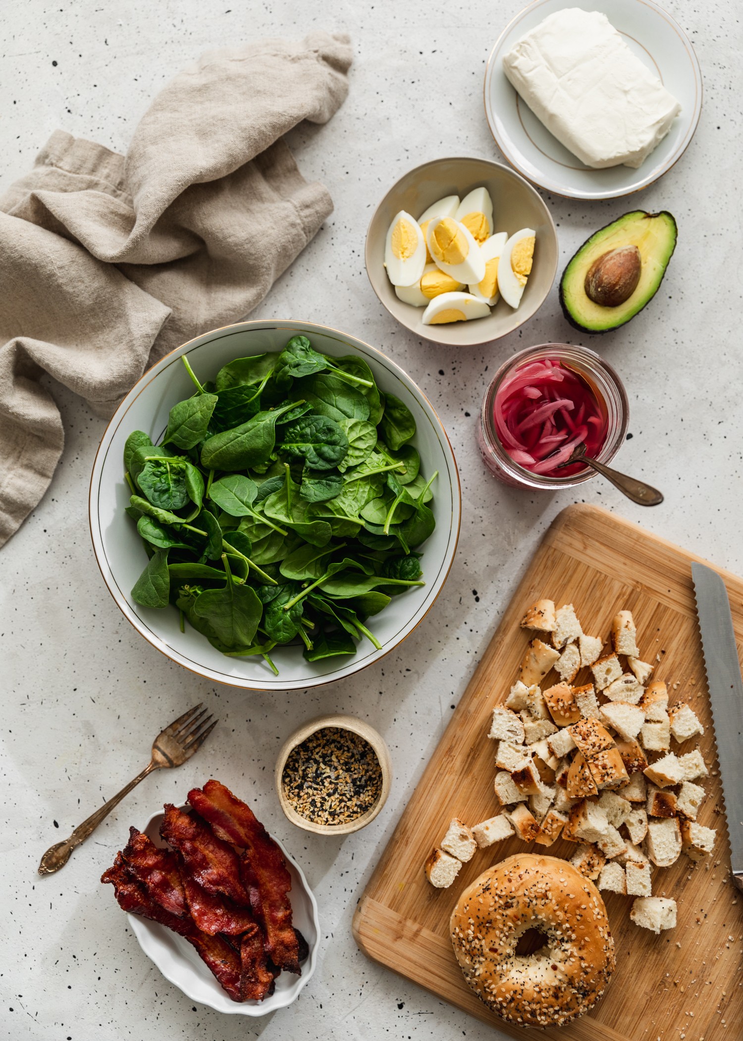 An overhead photo of salad ingredients including spinach, hard boiled eggs, bacon, and croutons on a grey counter.