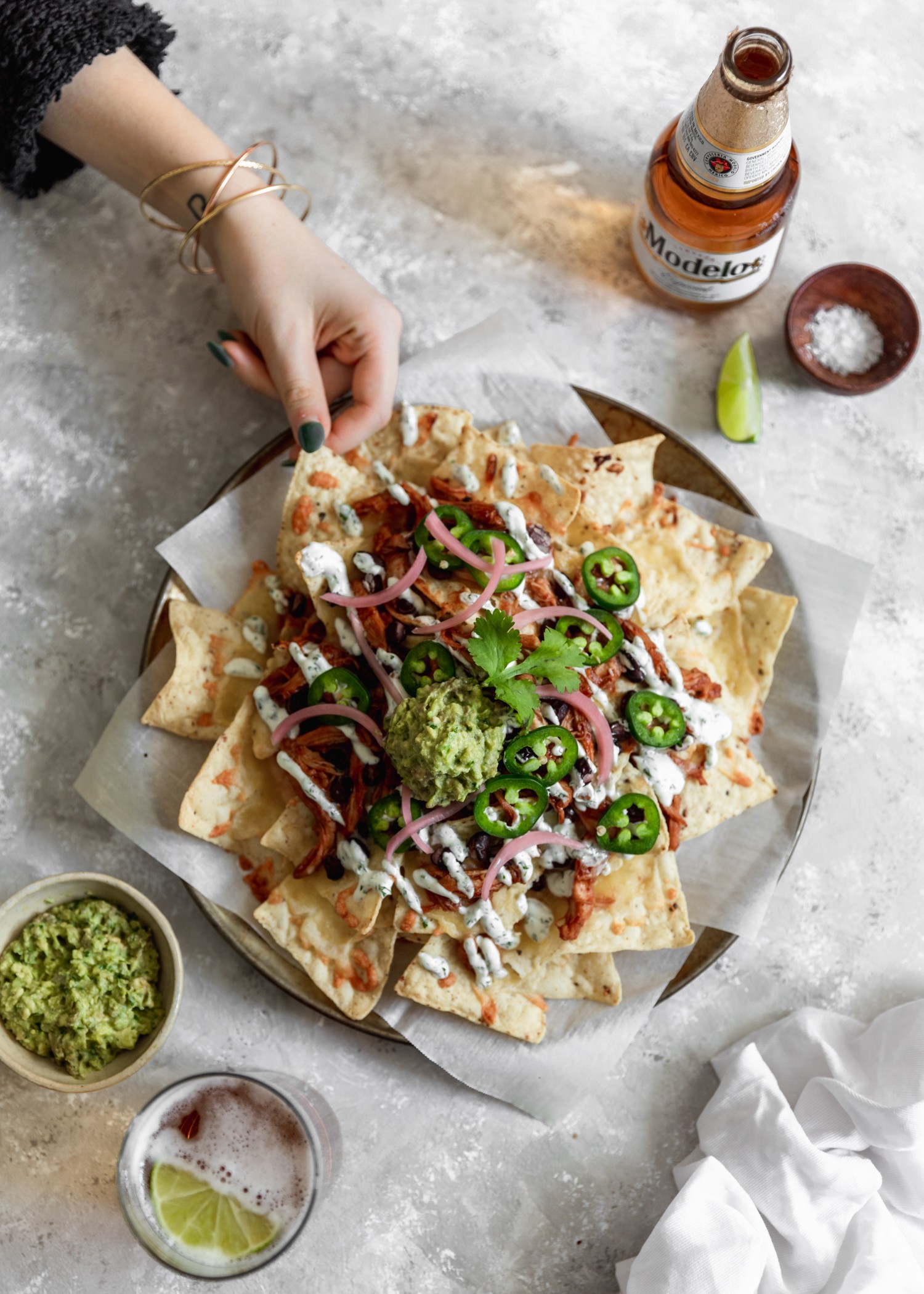 A woman's hand with bracelets and dark green nail polish reaching out to take a chip off of a plate of nachos on a grey table surrounded by ingredients.