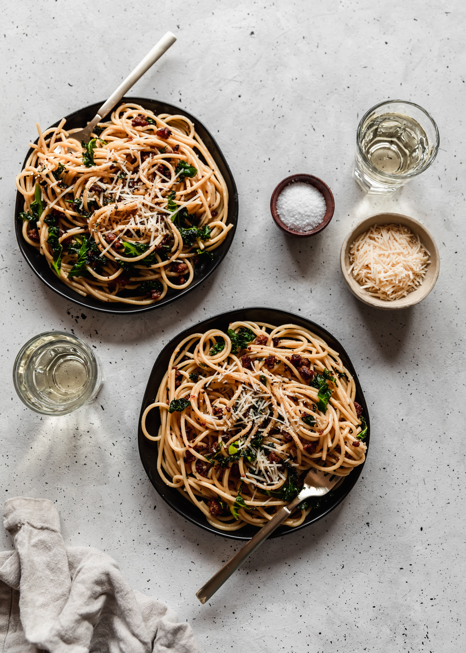 An overhead image of two plates of pasta with ground pork and greens on a grey table surrounded by white wine, a bowl of parmesan, a grey linen, and a small wood bowl with salt.