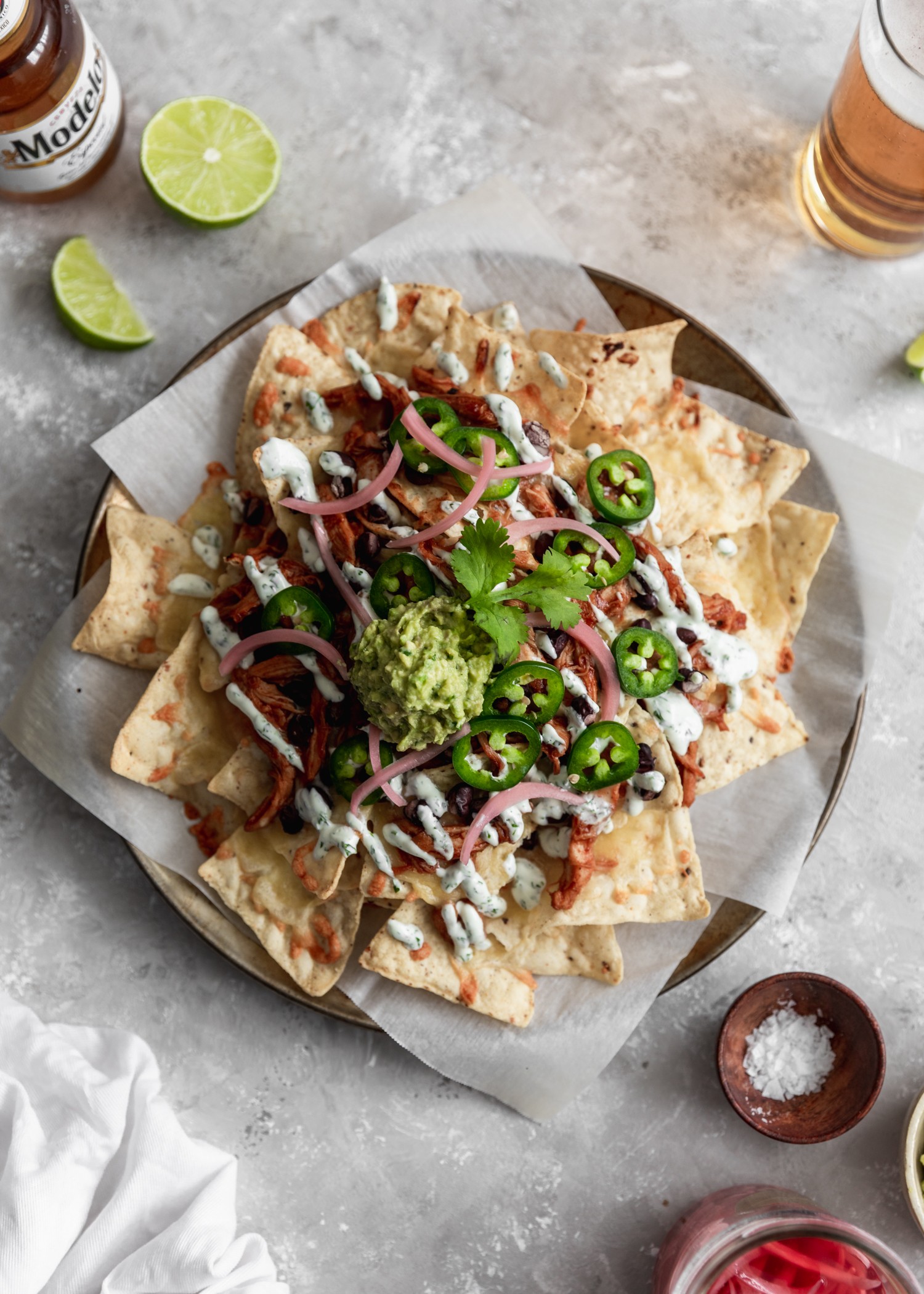 A closeup overhead image of a metal circle tray of BBQ chicken nachos on a grey counter surrounded by beer, lime wedges, pickled onions, and a wooden bowl of salt.