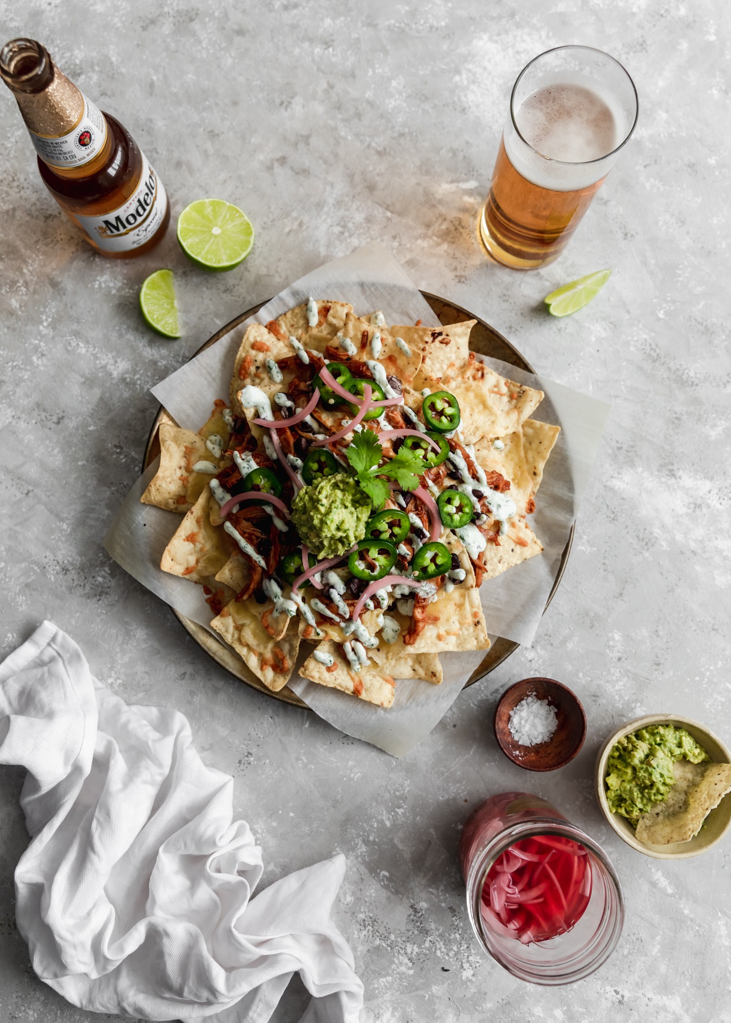 A bird's eye picture of a plate of tortilla chips topped with cheese, meat, and veggies on a grey counter surrounded by beer and a bowl of guacamole.