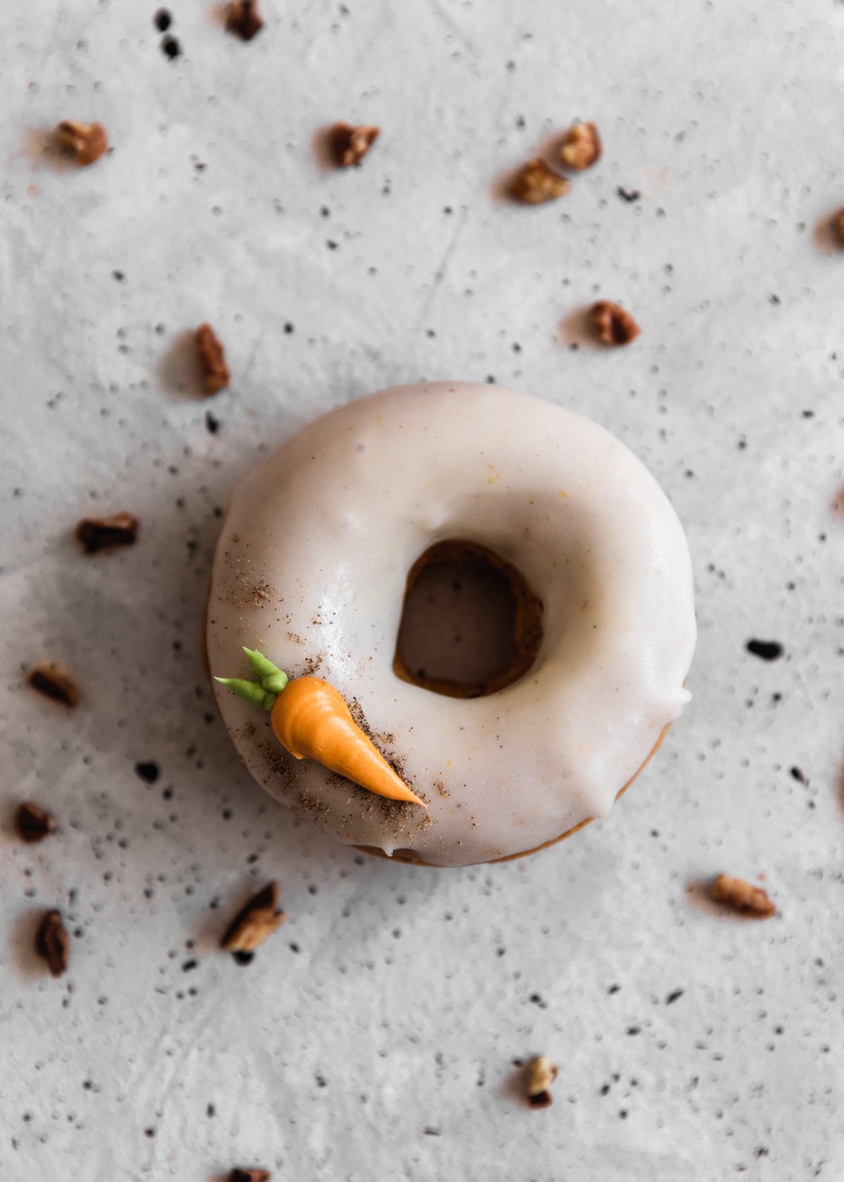 A closeup image of a carrot cake donut with white glaze and a frosting carrot on a grey table surrounded by pecans.