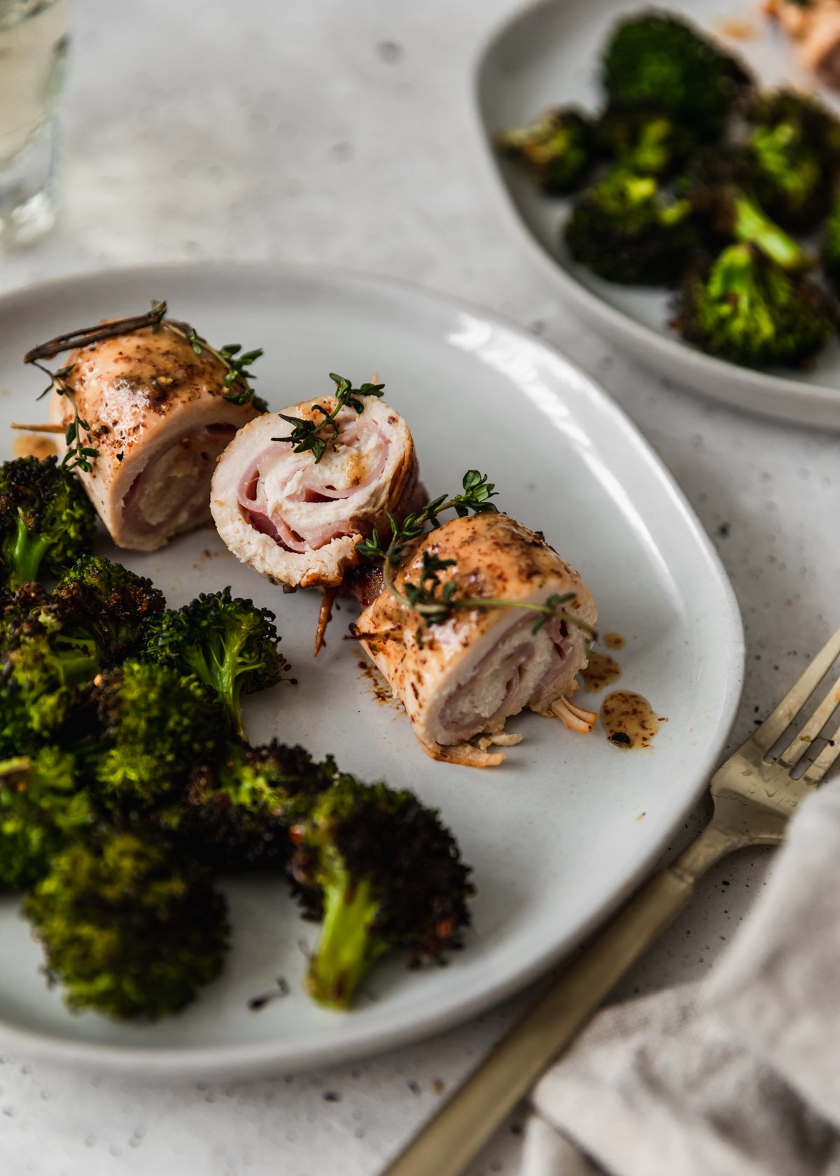A closeup side image of chicken cordon bleu rolls and broccoli on a white plate placed on a speckled white table.