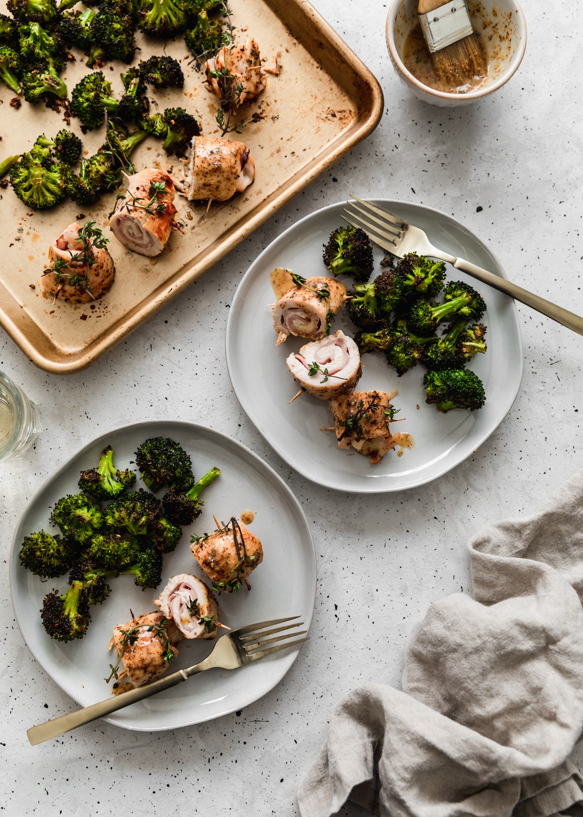 An overhead image of two white plates with sliced chicken cordon bleu and broccoli on a grey table with a gold sheet pan with more food in the left upper corner and a beige linen in the right lower corner.
