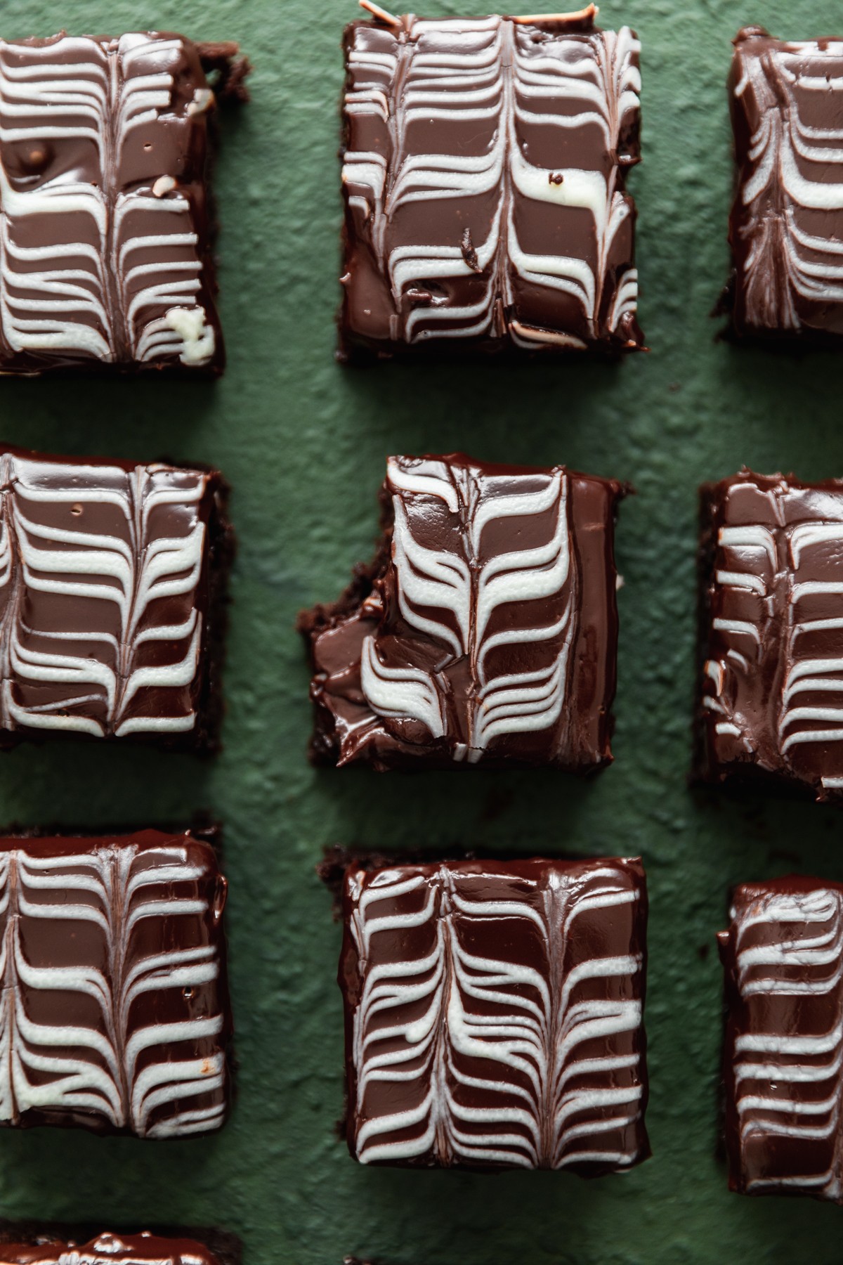 An overhead image of rows of brownies on a dark green background, with a bite taken out of the middle brownie.