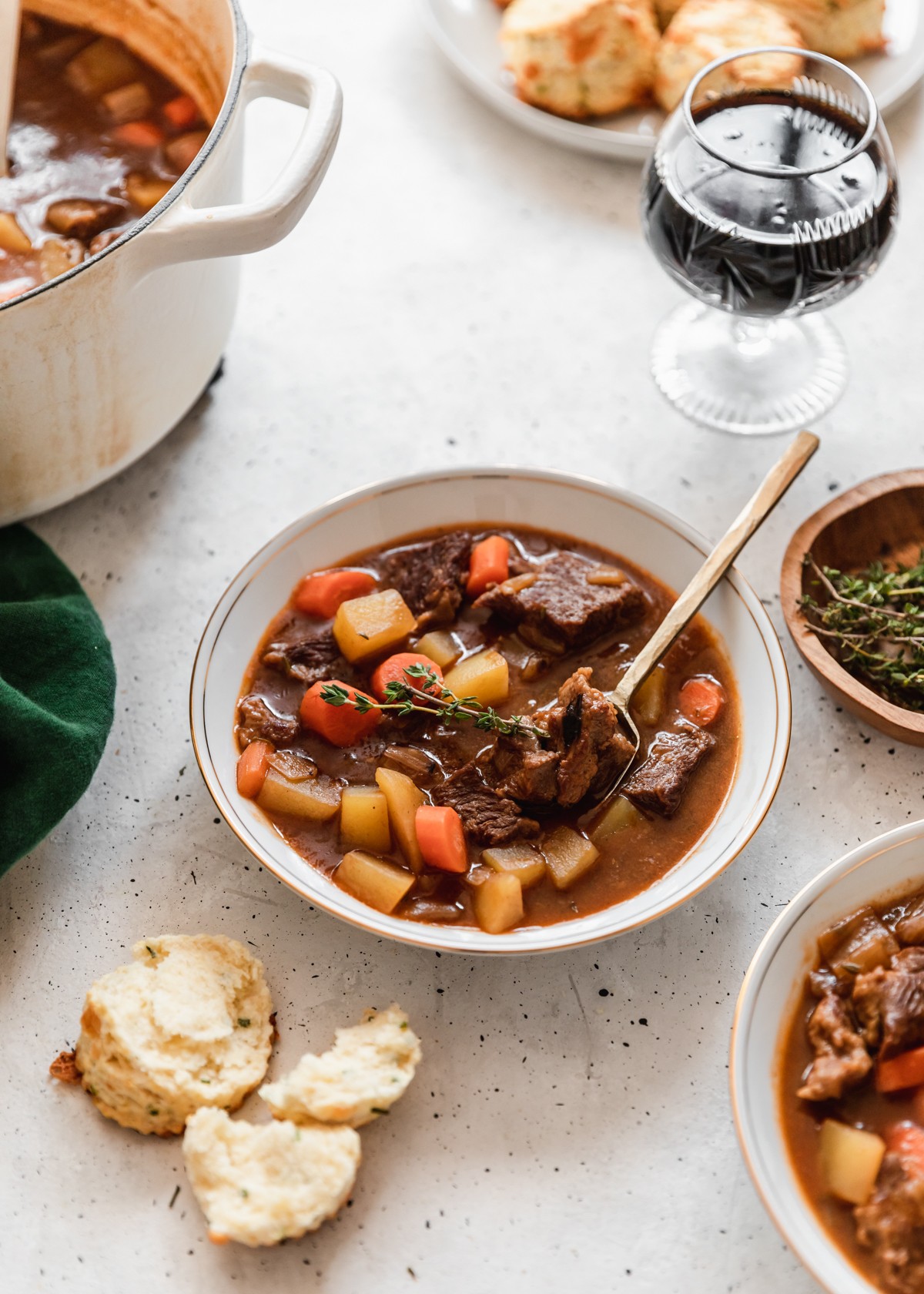 A side image off a white bowl of Irish beef stew on a white counter surrounded by another bowl of stew, wood bowl of thyme, glass of beer, and pot of soup.