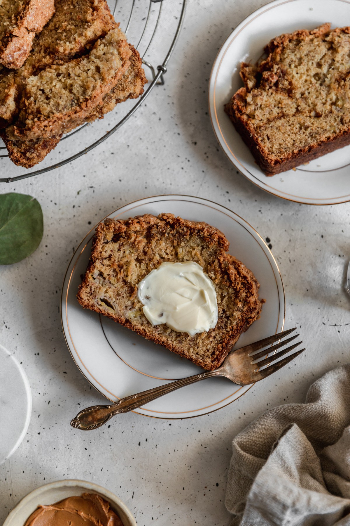 An overhead image of a slice of cookie butter banana bread topped with butter on a white plate placed on a grey counter and surrounded by coffee, a linen, and ingredients.