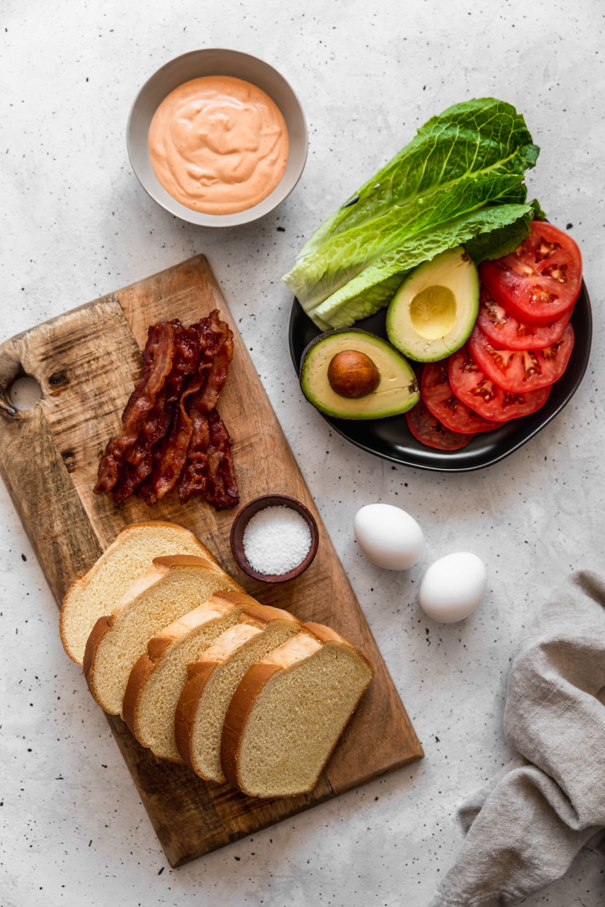 An overhead image of a black plate with lettuce, tomato slices, and a halved avocado on a grey table next to eggs, a wood board topped with bacon and bread, and a bowl of spicy mayo.