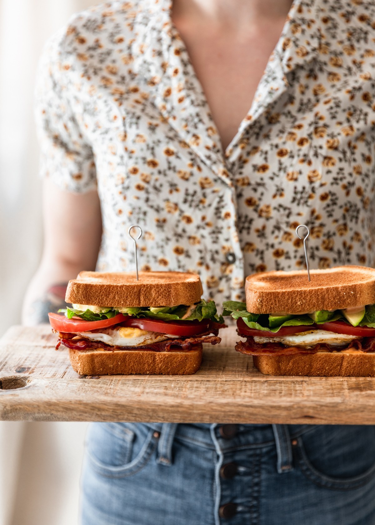 A side image of a woman wearing a white and yellow floral shirt and jeans holding a wooden board with two breakfast BLTs on top.