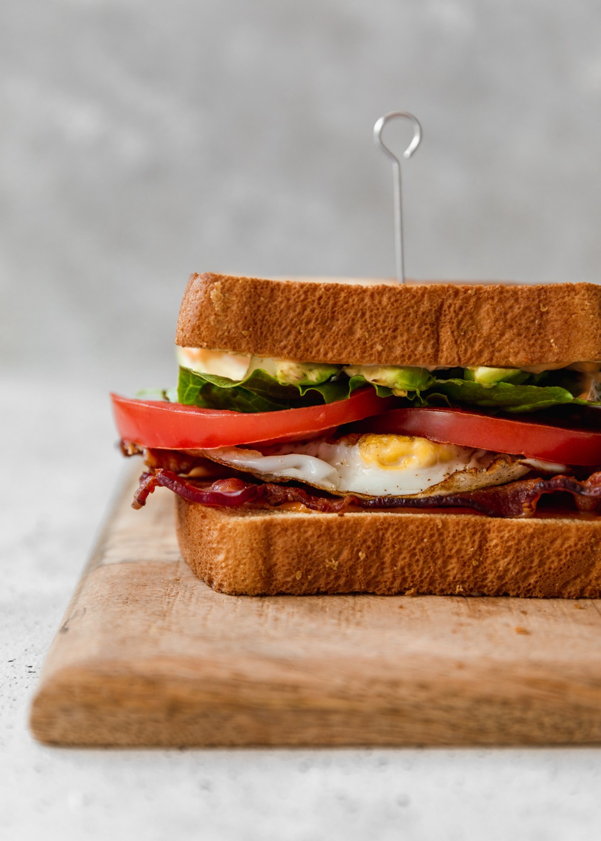 A very close up side image of a sandwich with bacon, lettuce, tomato, an egg, and avocado on a wood board with a grey background.