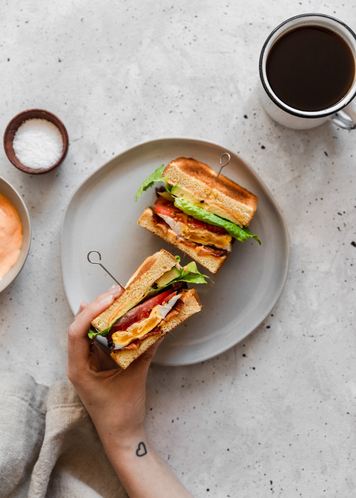 An overhead image of a woman's hands holding a breakfast BLT over a white plate on a grey table next to a cup of coffee and wooden bowl of salt.