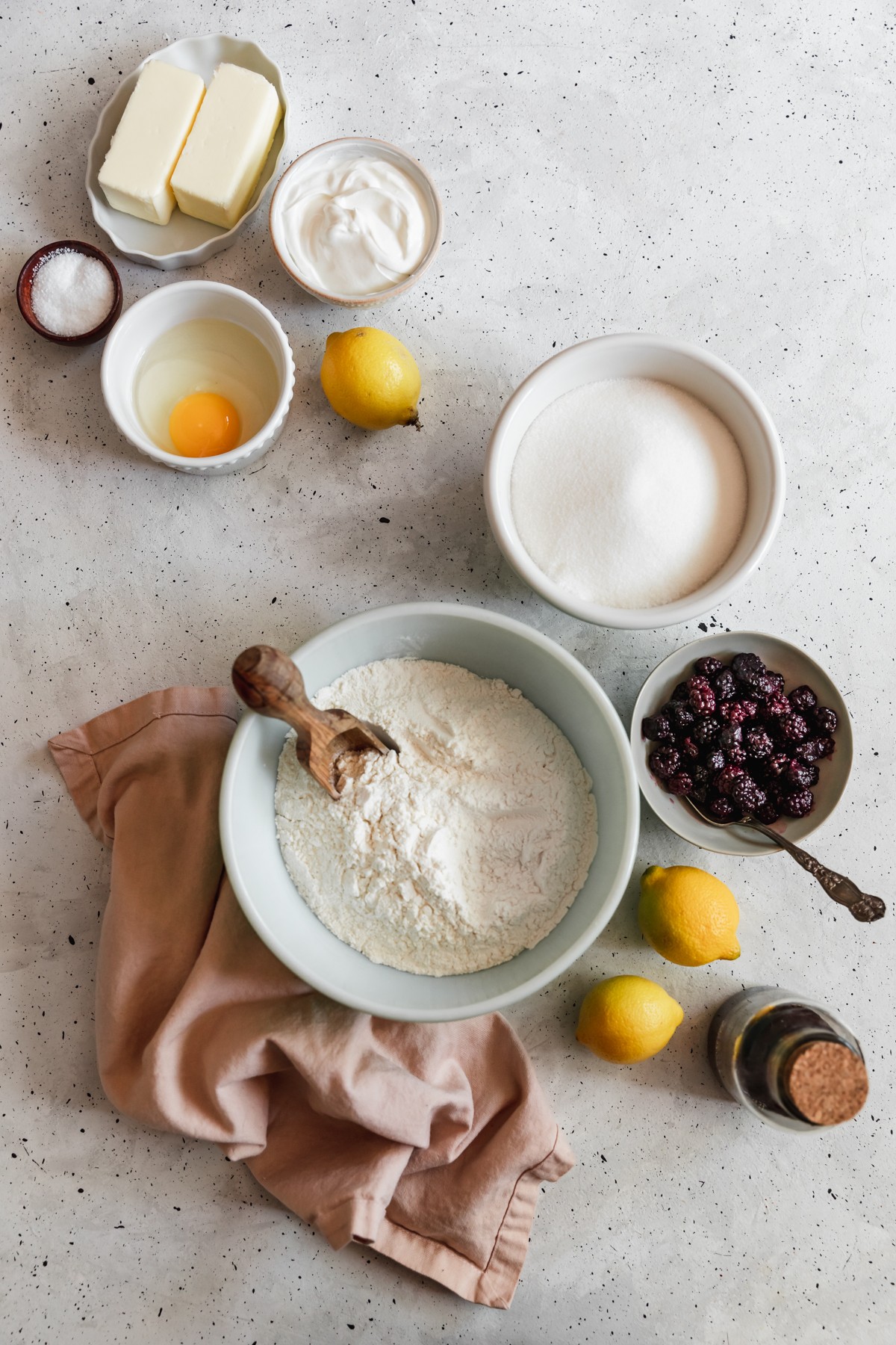 An overhead image of ingredients for sour cream lemon pound cake in various white and grey bowls on a white speckled table next to a bowl of blackberries, lemons, and a pink linen.
