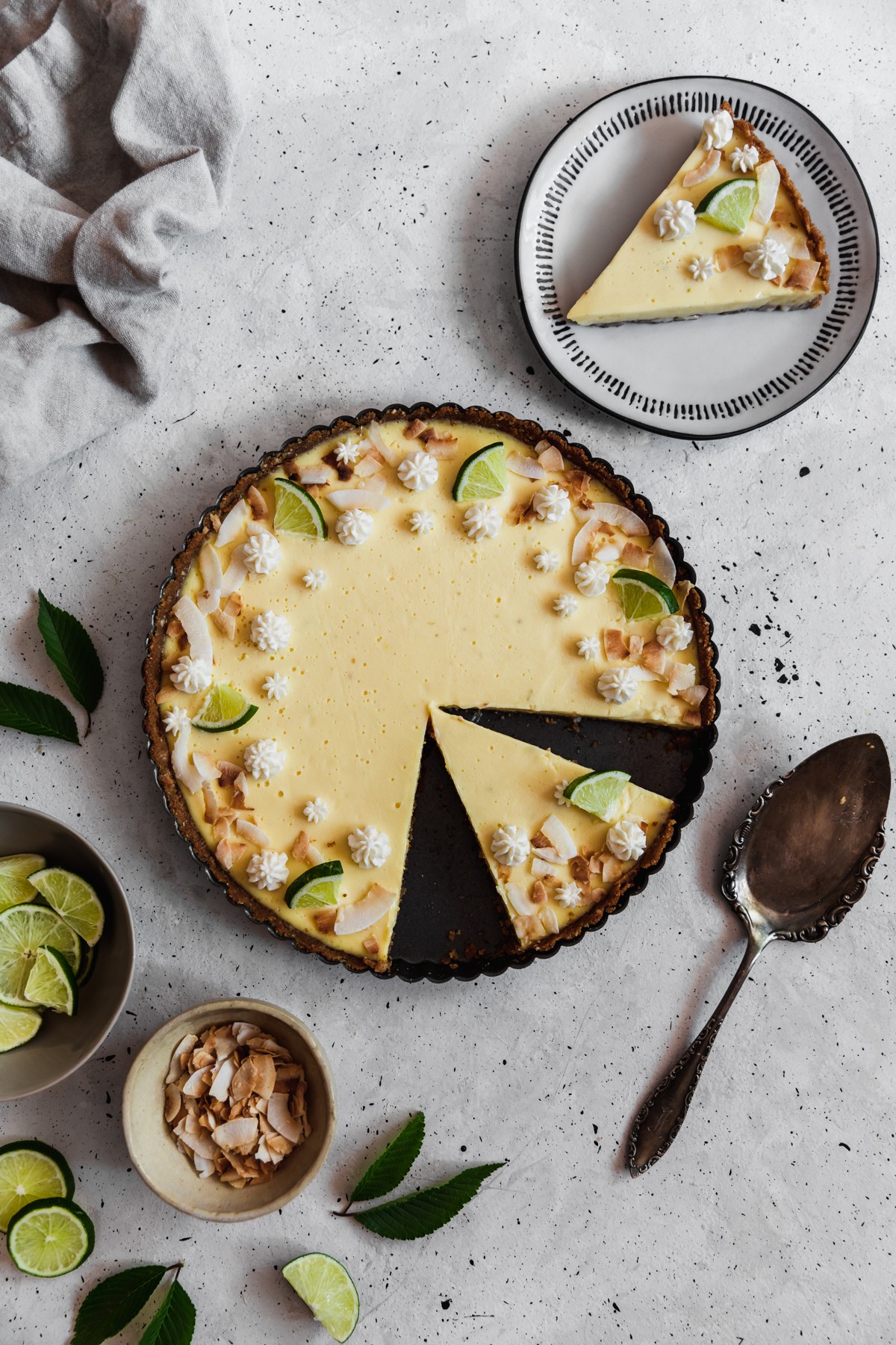 An overhead image of a sliced coconut key lime tart in a black pan on a grey table surrounded by a white plate topped with a tart slice, a beige linen, a vintage pie server, and a grey bowl of limes.