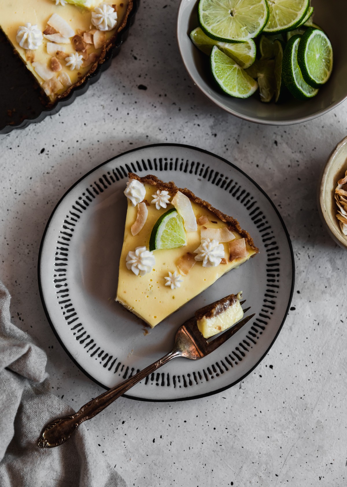 A closeup overhead image of a white plate topped with a slice of coconut key lime pie with a bite taken out of it on a grey table next to a cream bowl of toasted coconut and grey bowl of limes. 