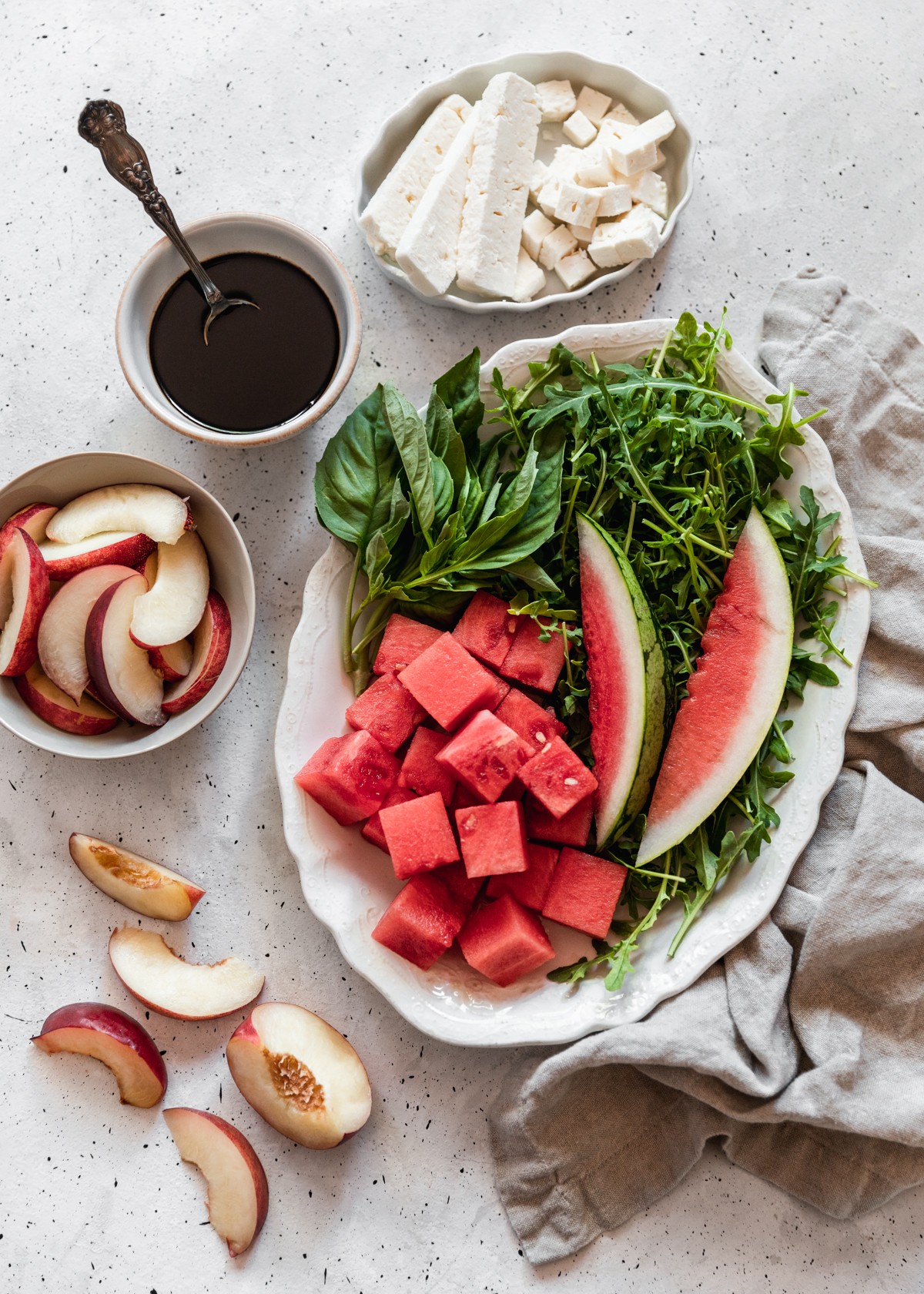 An overhead image of a white tray with cubed watermelon, arugula, and sprigs of basil on a speckled white table next to a beige linen, grey bowl of peaches, white bowl of balsamic, and oval white dish filled with feta cubes.