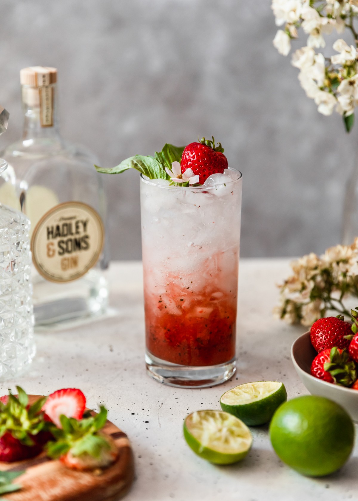 A side image of a strawberry gin smash garnished with a strawberry, basil, and white flower on a white table next to a grey bowl of strawberries, bar glasses, bottle of gin, and halved limes with a grey and white background.