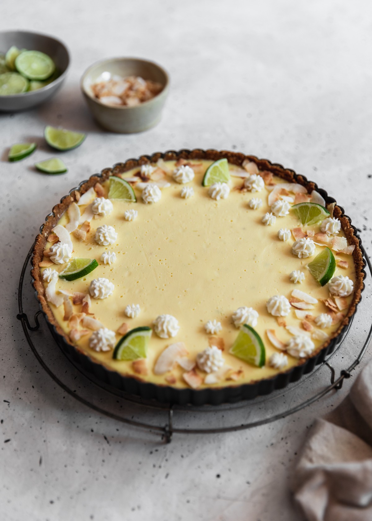 A side image of a coconut key lime pie on a cooling rack placed on a grey table next to a bowl of coconut flakes, bowl of limes, and grey linen.