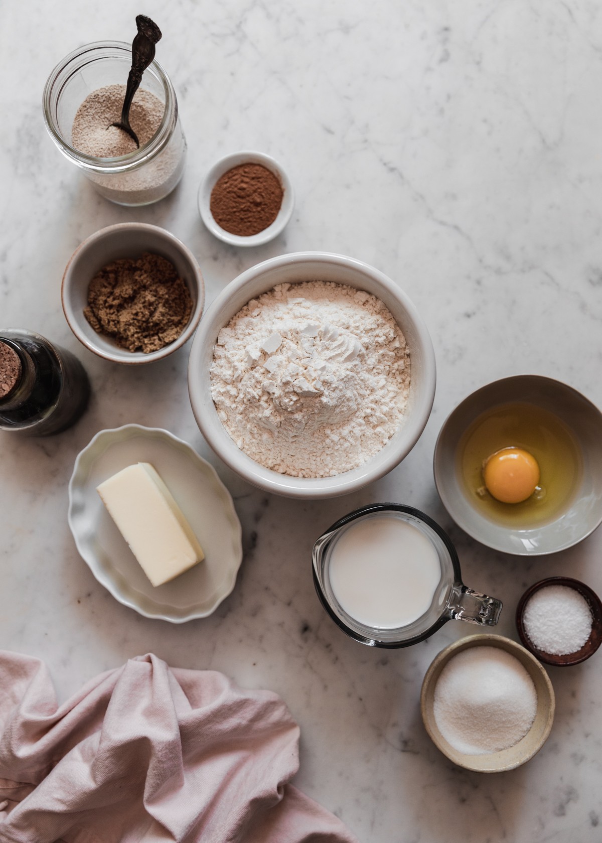 Various white and grey bowls of baking ingredients including flour, butter, brown sugar, and egg, salt, and vanilla on a marble counter next to a pink linen.