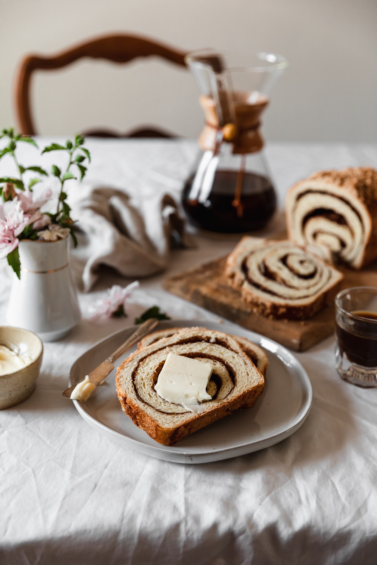 A white plate with two slices of cinnamon swirl bread topped with a smear of butter next to a wood board with a loaf of bread, a white vase of pink flowers, a cream-colored bowl of butter, a Chemex, and a beige linen on a white table cloth with a wooden chair in the background.
