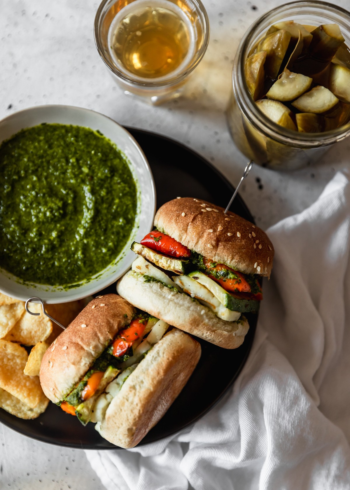 A closeup overhead image of two vegetable and halloumi sandwiches on a black plate next to a grey bowl of herb sauce, a jar of pickles, and a glass of beer. The plate is resting on a white linen with a light grey table as the background.