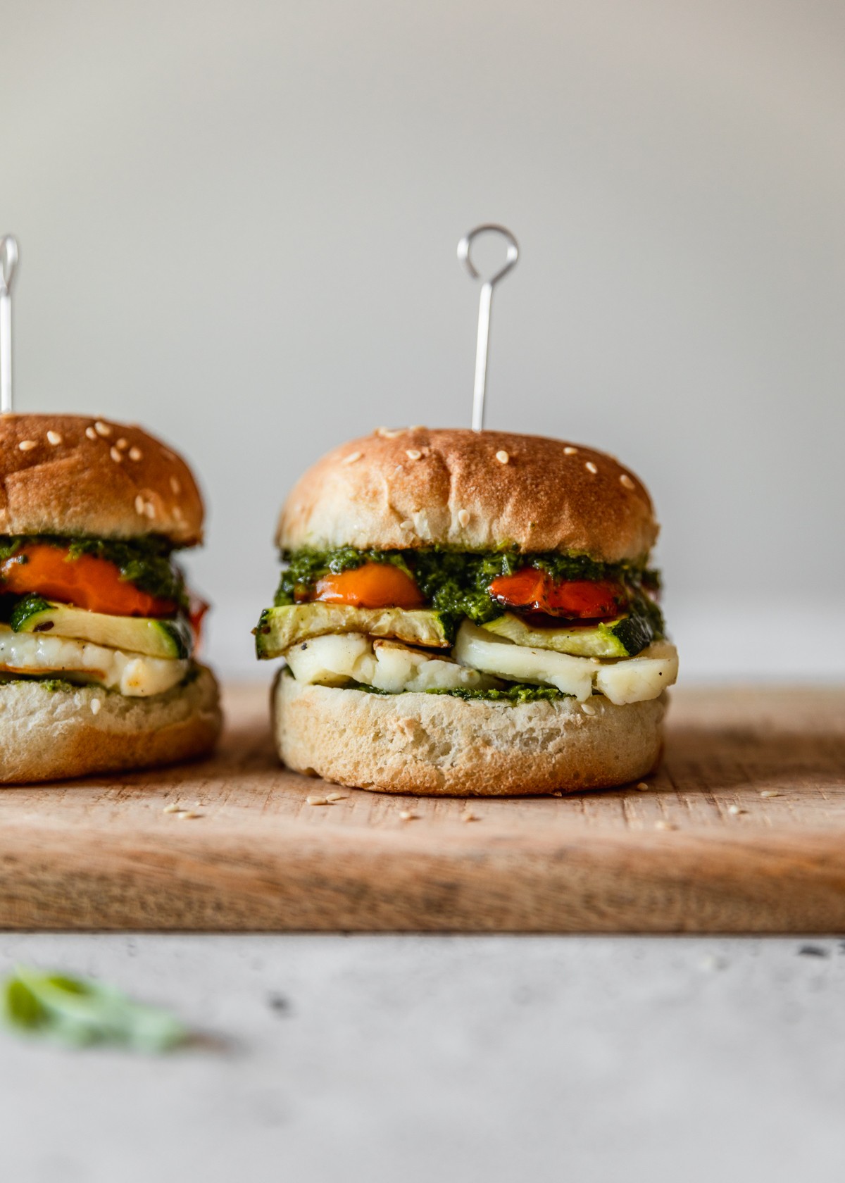 A closeup side image of a halloumi sandwich with veggies and herb sauce topped with a metal pick next to another sandwich on a wood board with a white background.