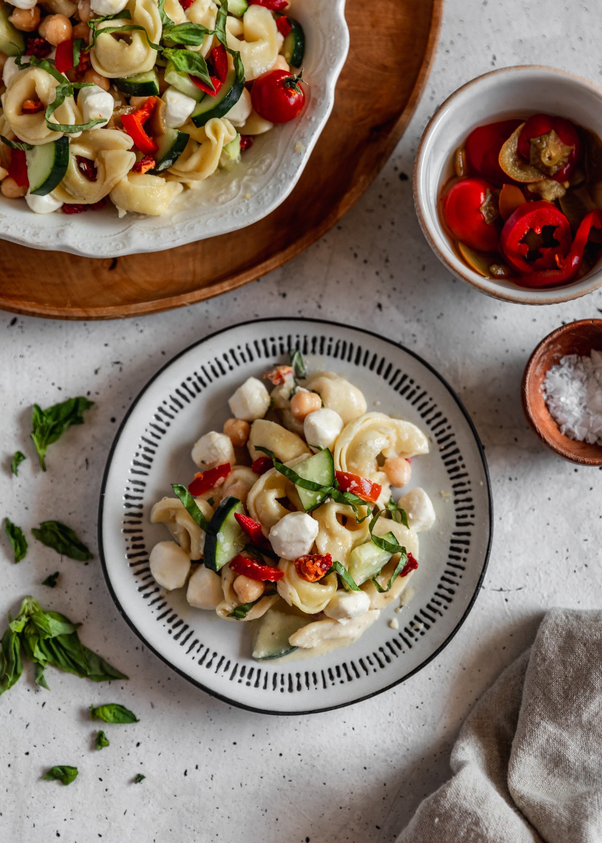 An overhead image of a white plate with a black rim topped with antipasto tortellini pasta salad next to a white bowl of peppers, wood bowl of salt, beige linen, and basil on a grey counter. In the left upper corner is part of a white plate topped with more pasta salad.