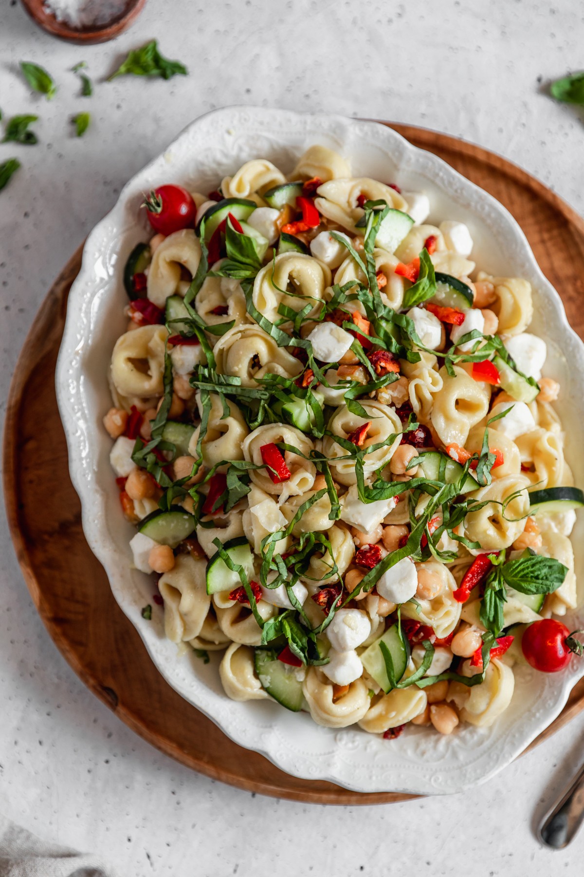 A closeup overhead image of a white oval plate topped with tortellini salad with veggies, Mozzarella, and basil placed on a wood tray next to a beige linen and basil on a grey speckled table.