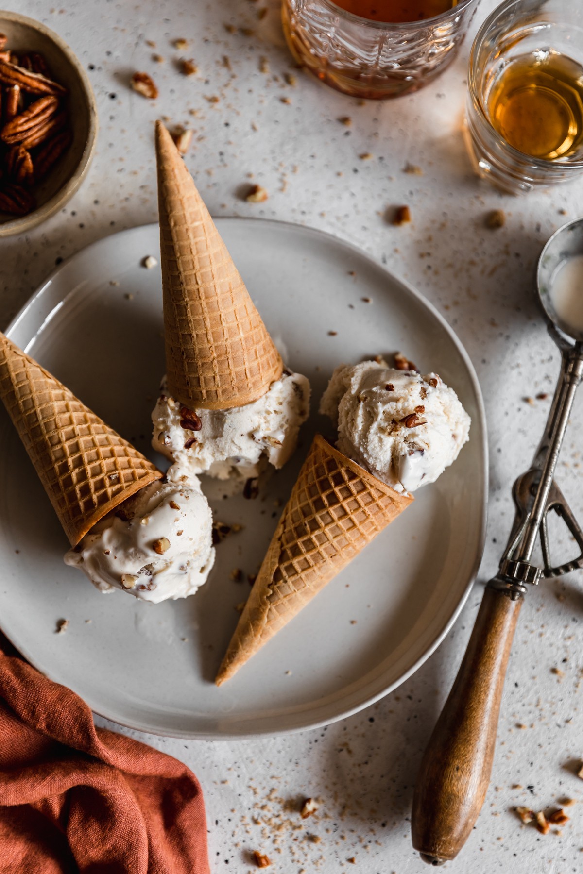 A closeup overhead image of three scoops of bourbon butter pecan ice cream with cones on a white plate next to a vintage ice cream scoop, two glasses of whiskey, and burnt orange linen. The plate is on a white speckled table dusted with chopped pecans.