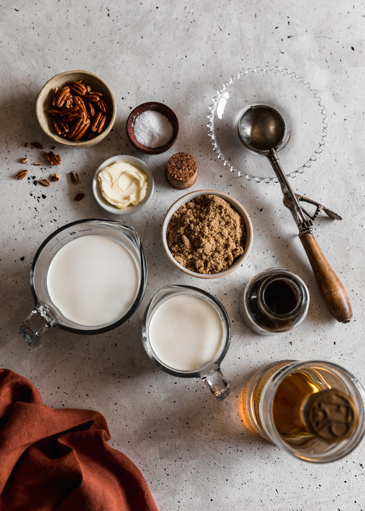 Two glass measuring cups with heavy cream and half-and half, a white bowl of brown sugar, a bottle of vanilla, a bottle of bourbon, and bowls of butter, salt, and pecans on a white speckled table. Next to the ingredients is a vintage ice cream scoop and dark orange linen.