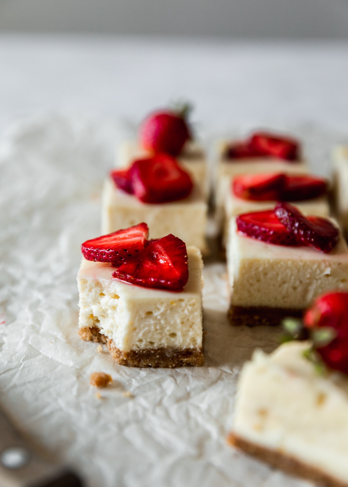 A closeup image of a strawberry cheesecake bar with a bite taken out of it on white parchment paper surrounded by rows of more bars.